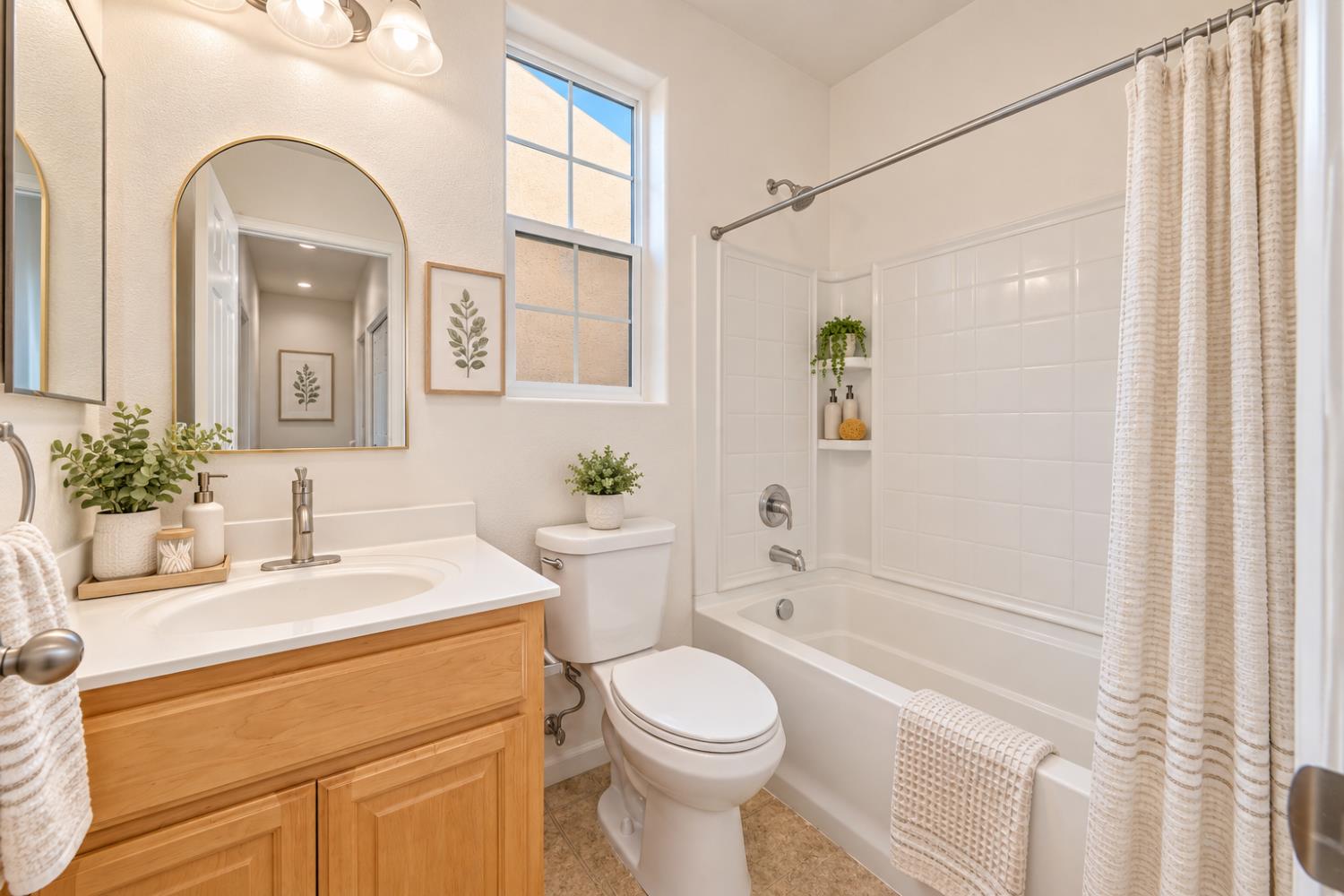 Bright bathroom with a light wood vanity, arched mirror, and white tub with tiled surround.