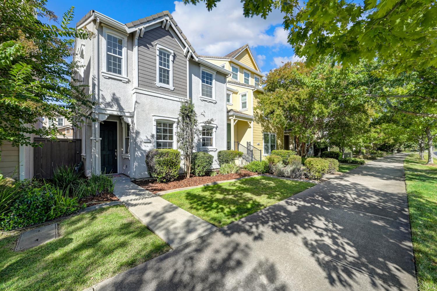 Gray two-story home with white trim and a dark front door beside a tree-lined sidewalk.