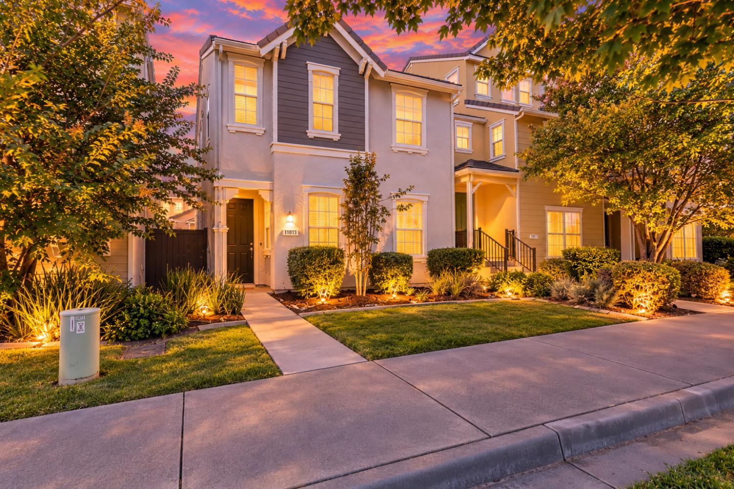 Two-story townhouse with a dark gray gabled facade and warmly lit landscaping at dusk.