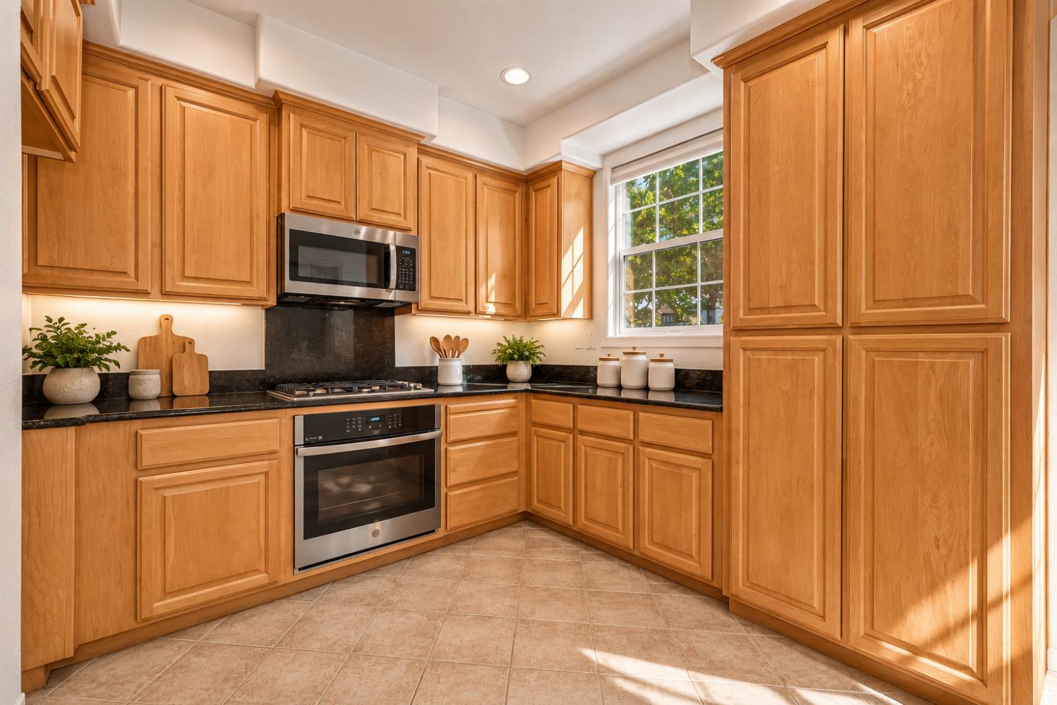 Kitchen with warm wood cabinets, dark countertops, stainless steel appliances, and a sunlit window.