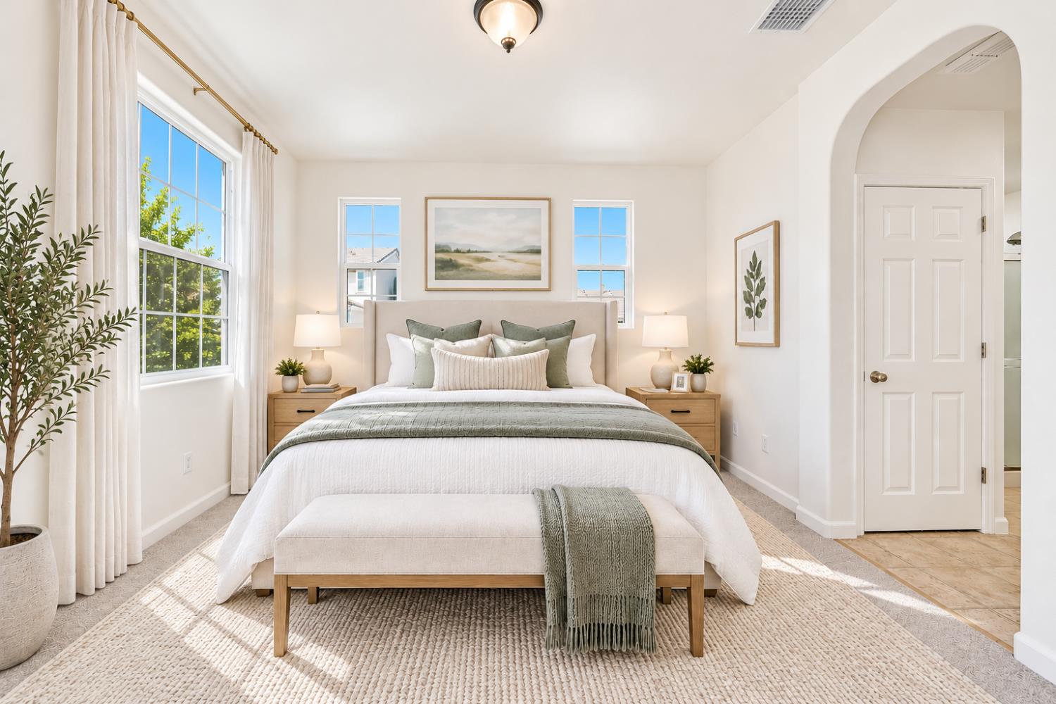 Staged bedroom with a large bed, upholstered bench, and tall curtained windows.