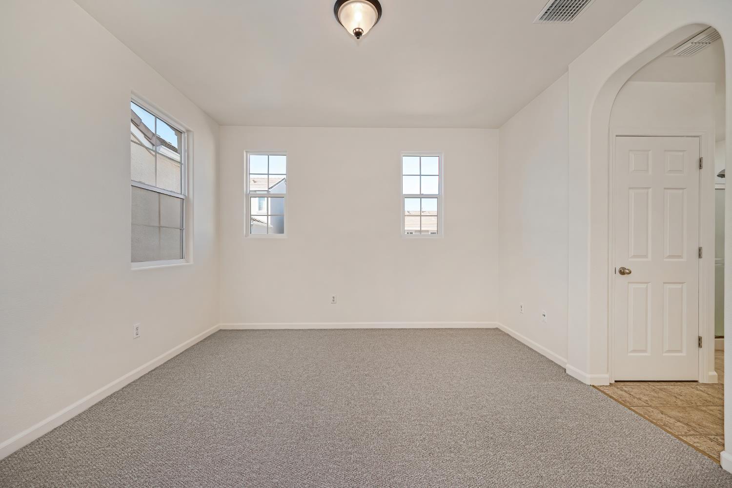 Empty carpeted room with three windows and an arched doorway to a tiled area.