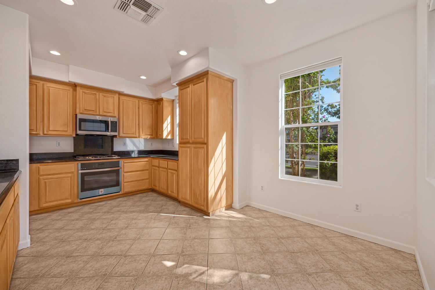 Vacant kitchen with honey-colored cabinets, dark countertops, built-in appliances, and a large sunlit window.