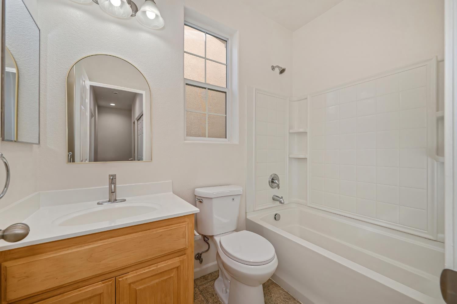 Bathroom with tiled tub and shower, wooden vanity, arched mirror, and tall window.