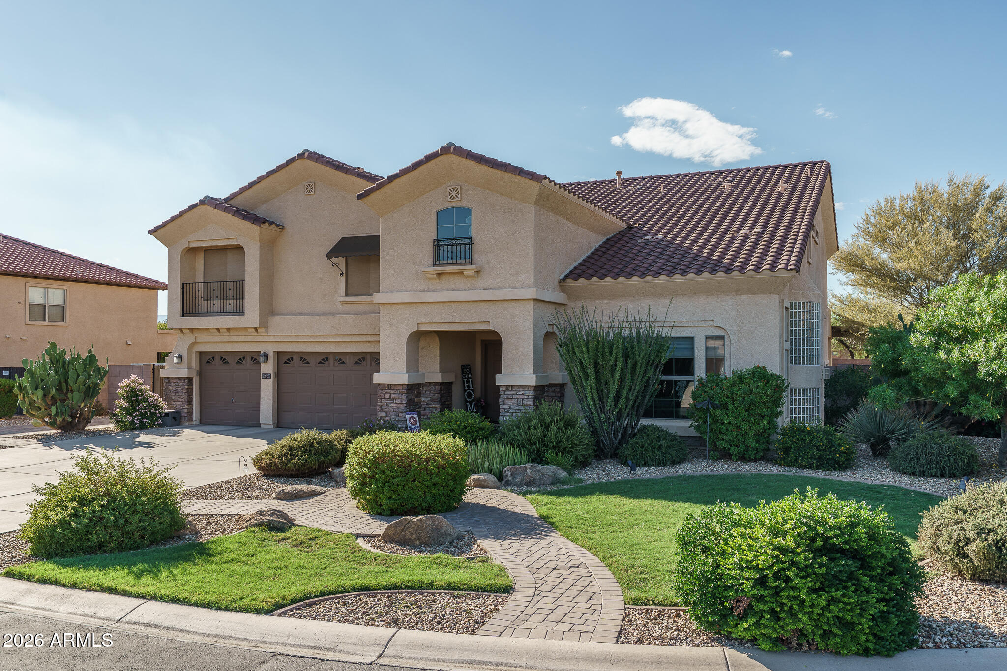 Two-story stucco home with a tiled roof, double garage, balcony, and curved walkway through a landscaped yard.
