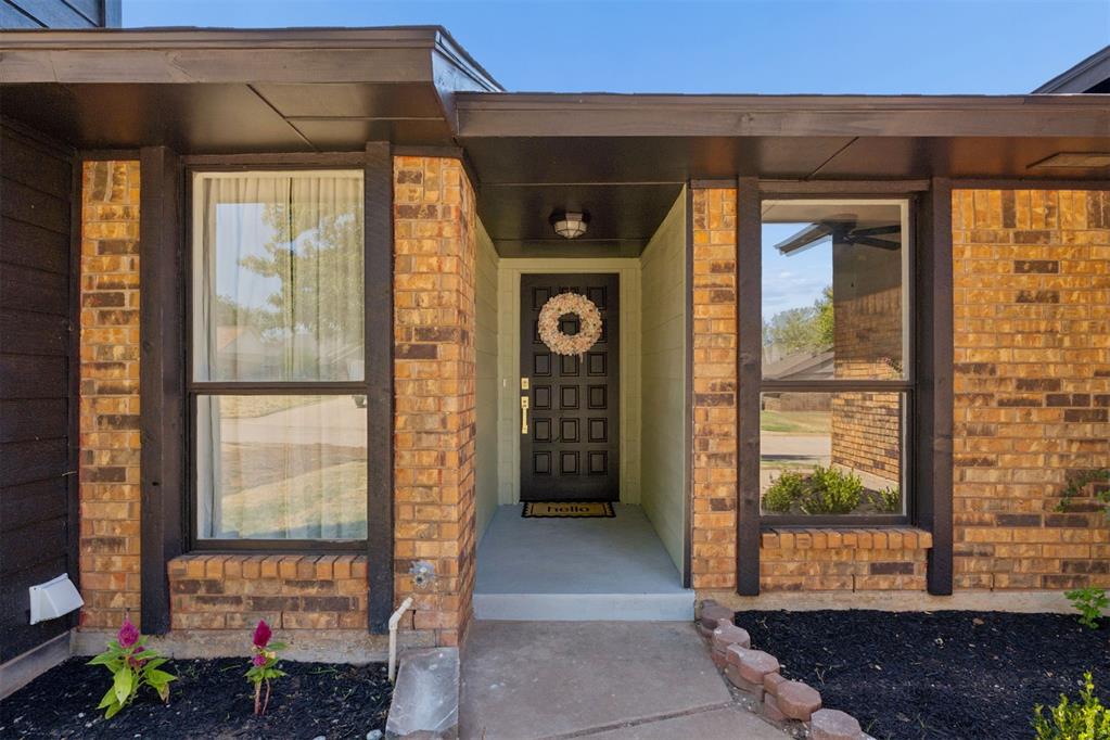 Brick home entrance with a recessed dark door, wreath, flanking windows, and flower beds.