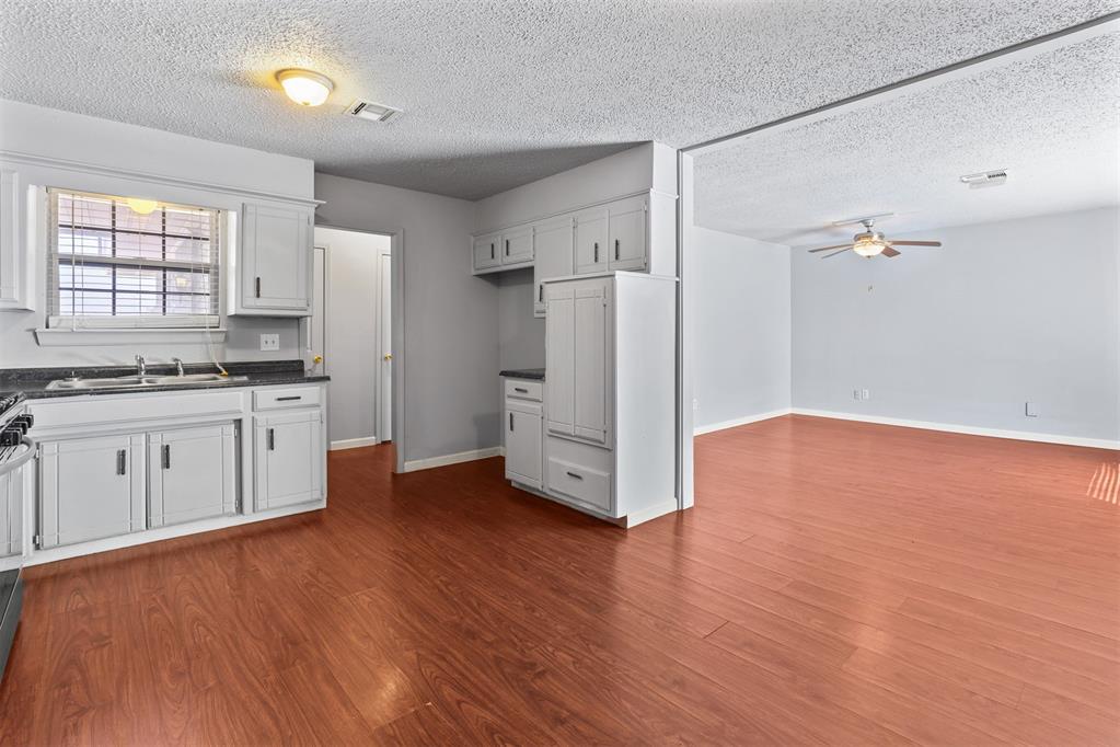 Kitchen opening into an unfurnished living area with white cabinets and reddish wood-look flooring.