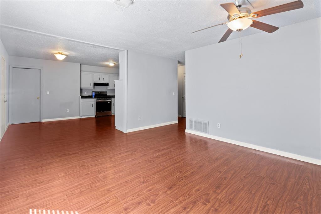 Empty living room with wood-look floors and a ceiling fan, opening into a kitchen.