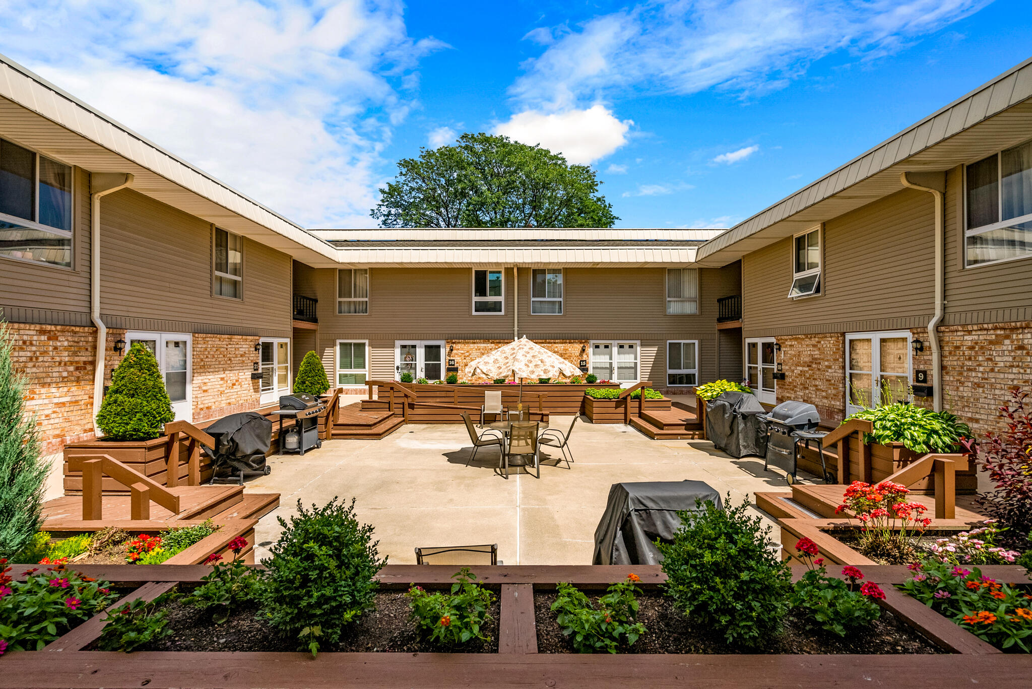 Apartment courtyard with raised flower beds, patio seating, grills, and a central umbrella.