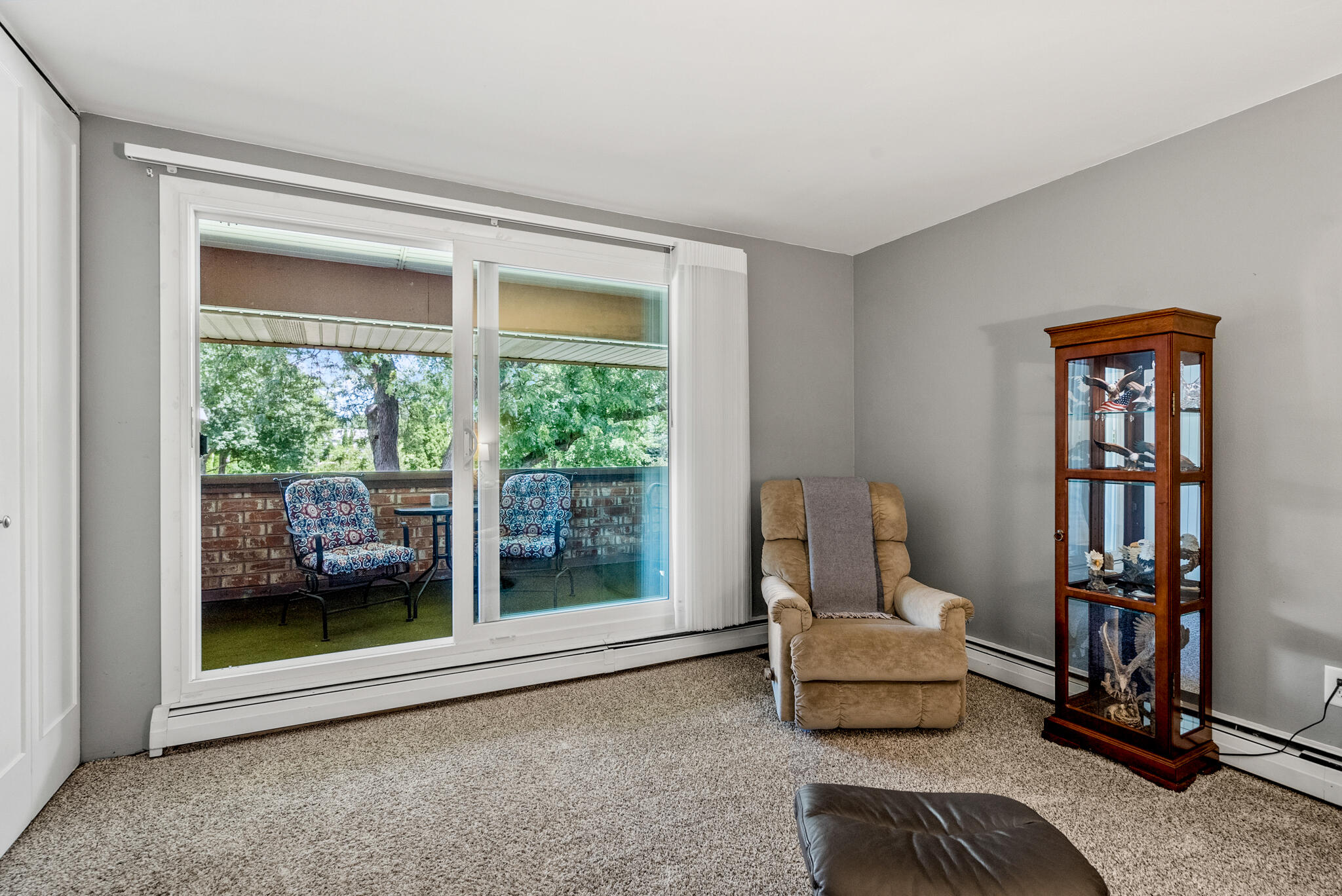 Living room with a tan recliner and display cabinet beside a sliding door to a tree-lined balcony.