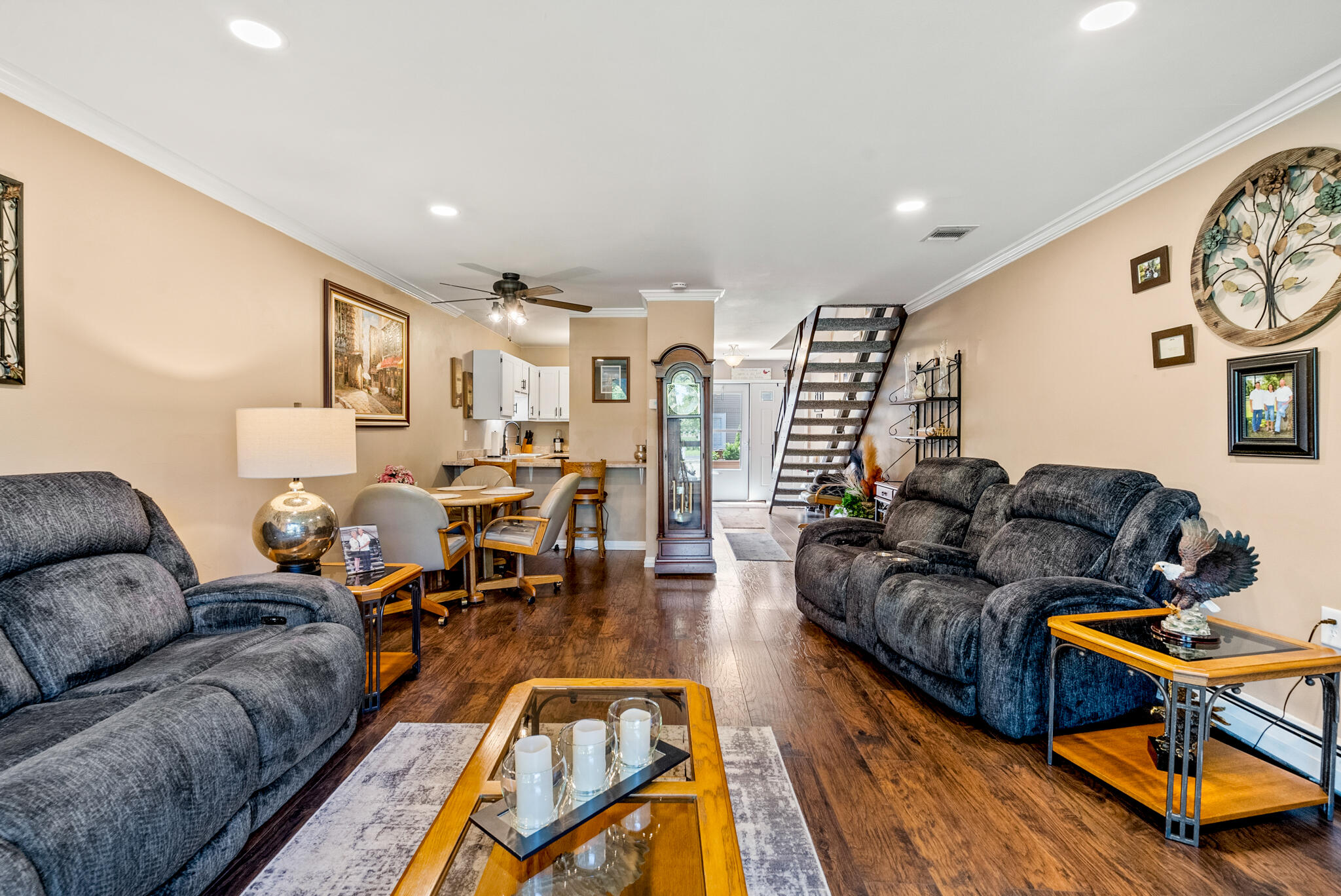 Open-plan living room with gray reclining sofas, hardwood floors, and a staircase beside the dining area.