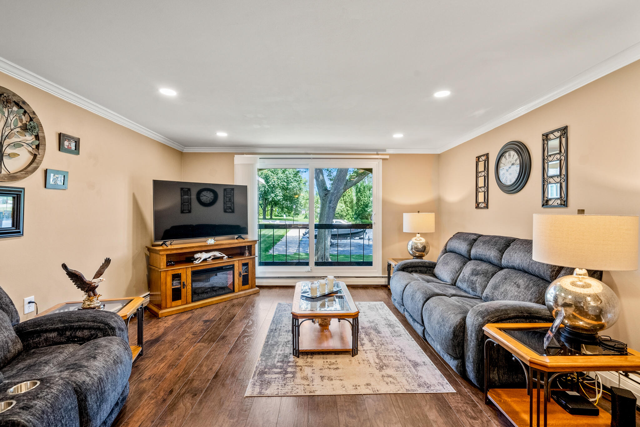 Furnished living room with gray reclining sofas and sliding glass doors overlooking a leafy yard.