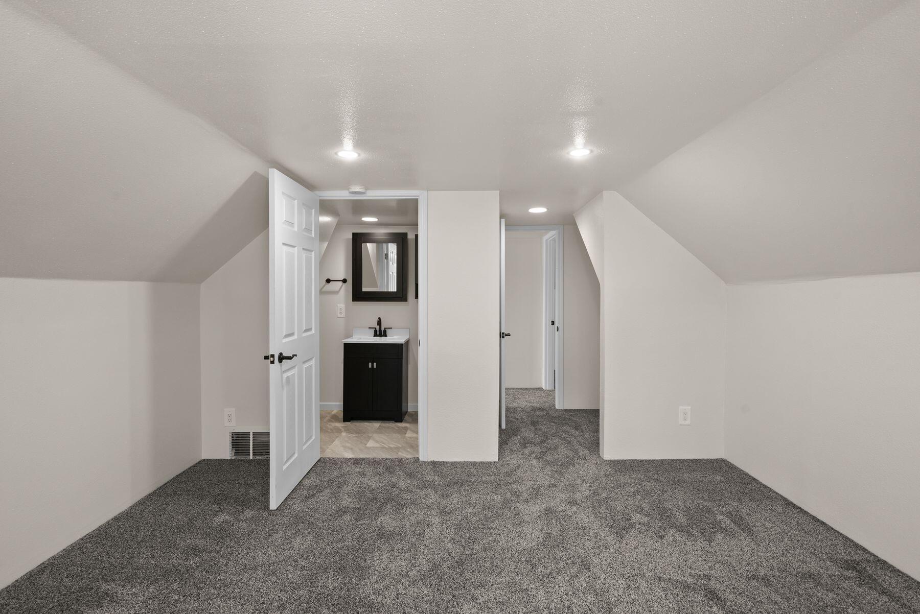 Finished attic room with gray carpet and recessed lights, looking toward an open bathroom door.