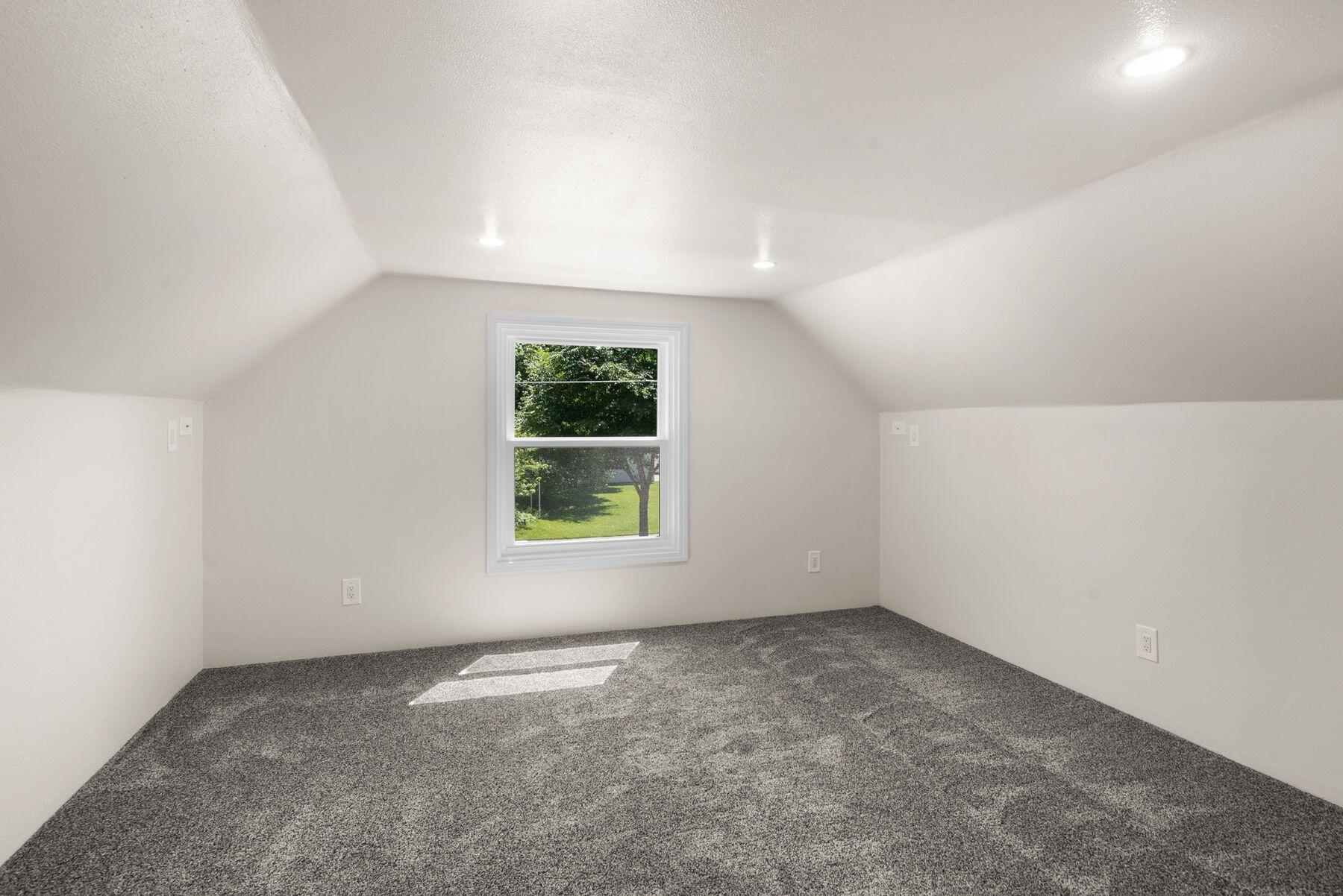 Vacant carpeted room with sloped ceilings and a window overlooking a green yard.
