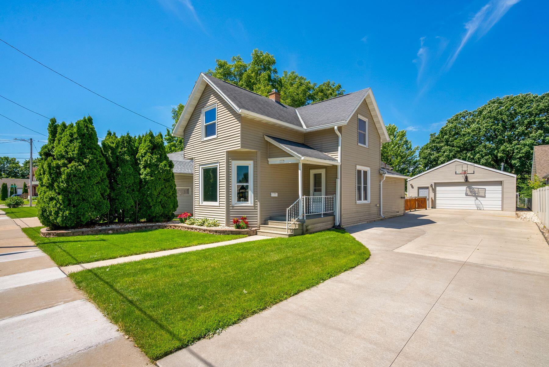 Two-story beige house with a covered porch, landscaped lawn, and driveway to a detached garage.