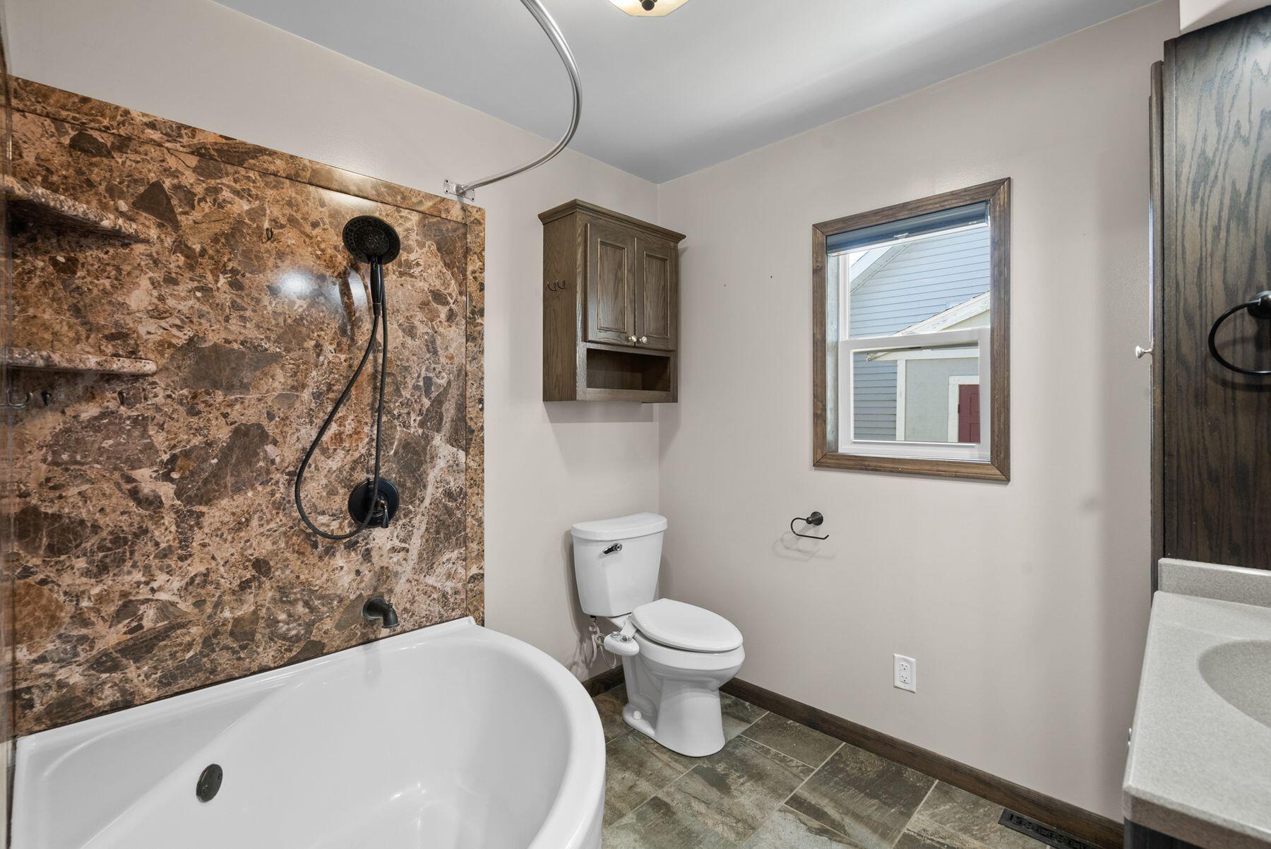 Bathroom with a marble-look shower surround, white bathtub, toilet, and wood-framed window.