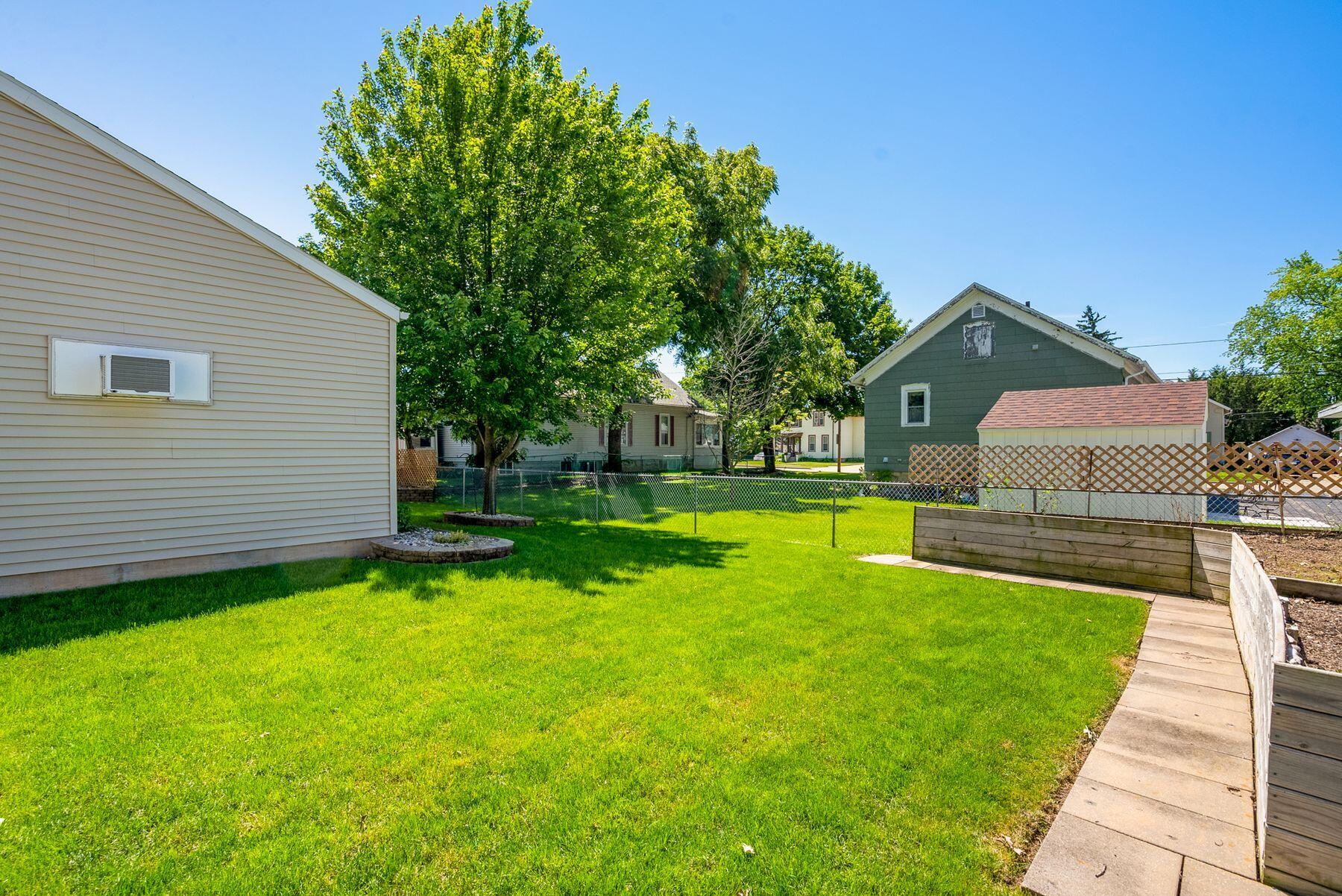 Backyard with a broad lawn, mature shade trees, raised garden beds, and a concrete walkway.