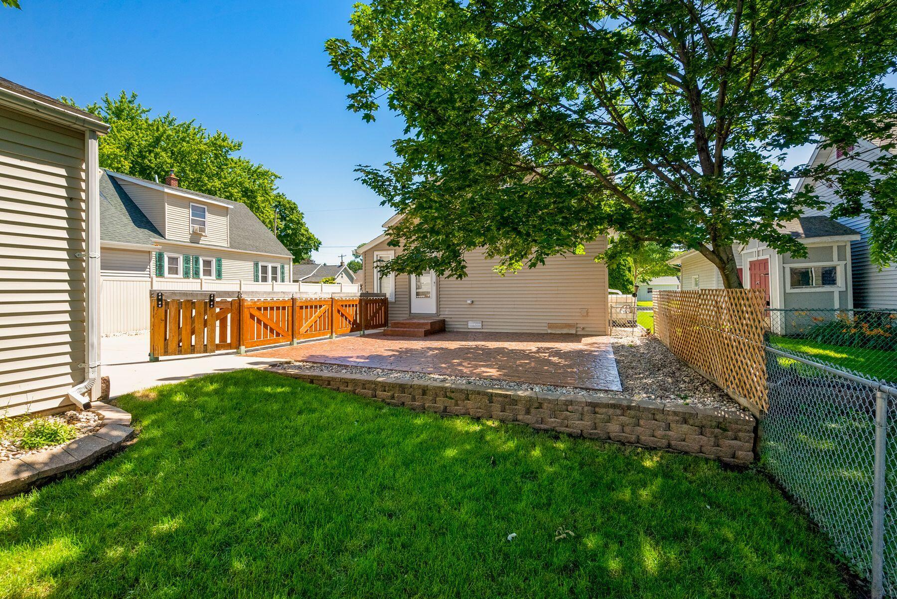 Backyard patio bordered by wooden railing and fencing, with a large shade tree and green lawn.