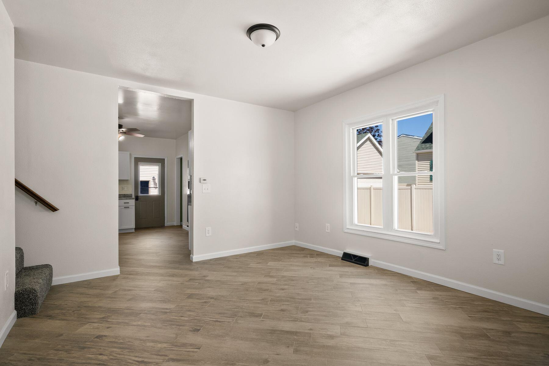 Empty living room with wood-look tile flooring, a large window, and an opening to the kitchen.