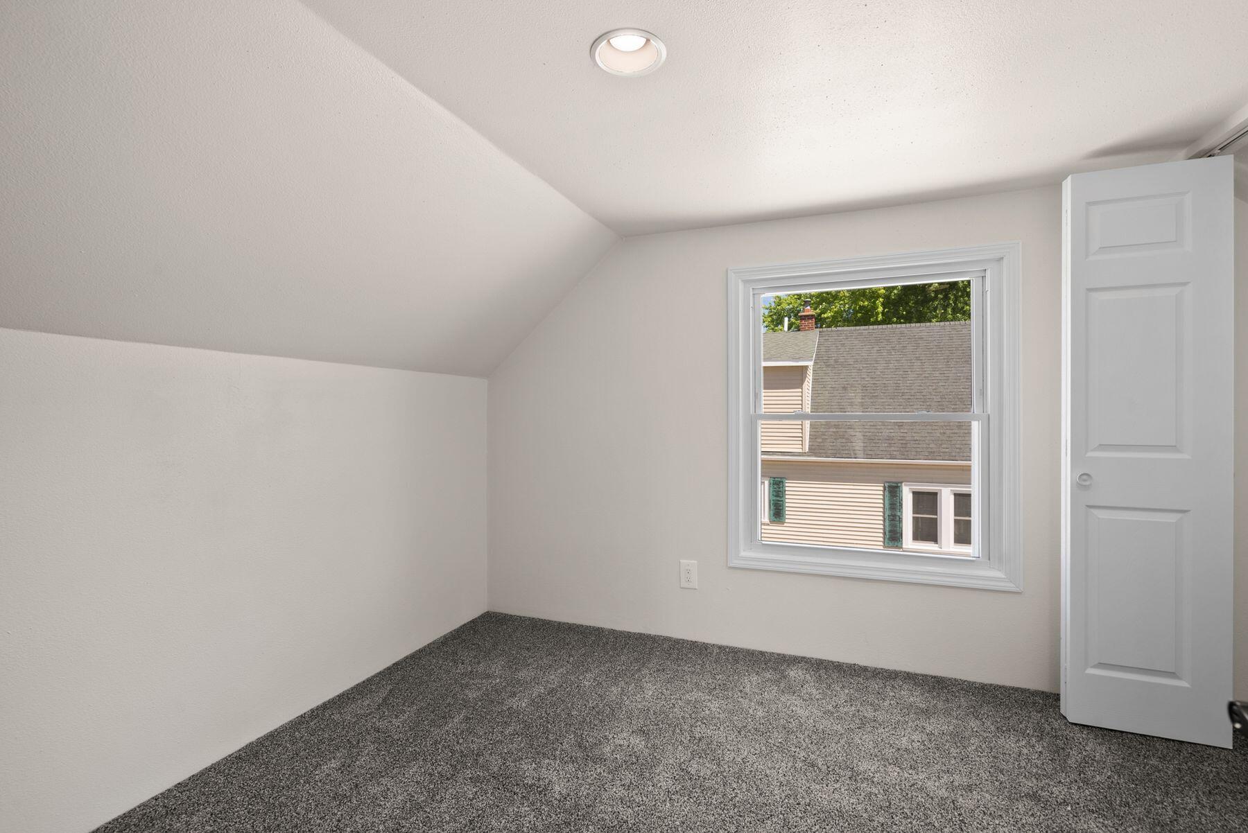 Vacant bedroom with gray carpet, a sloped ceiling, and a window facing a neighboring home.