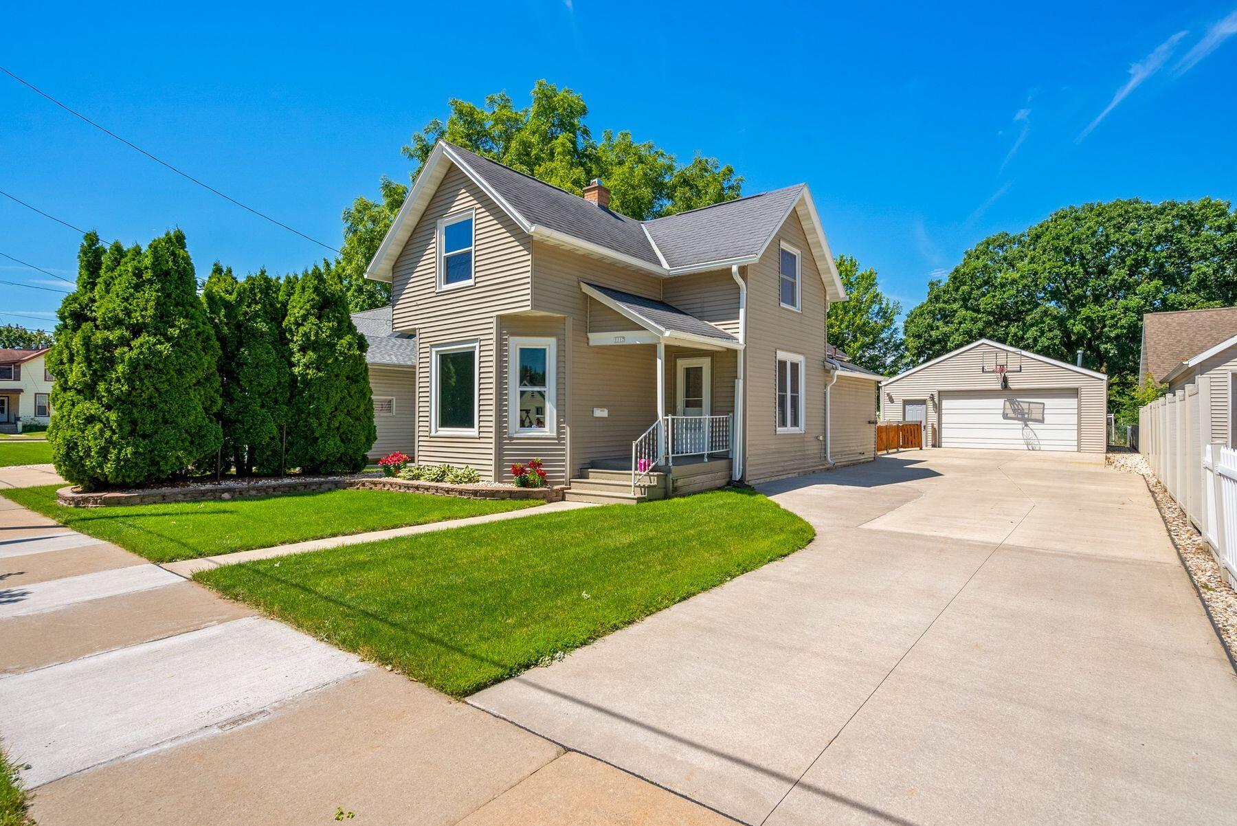 Two-story tan house with a covered porch, green lawn, and driveway to a detached garage.