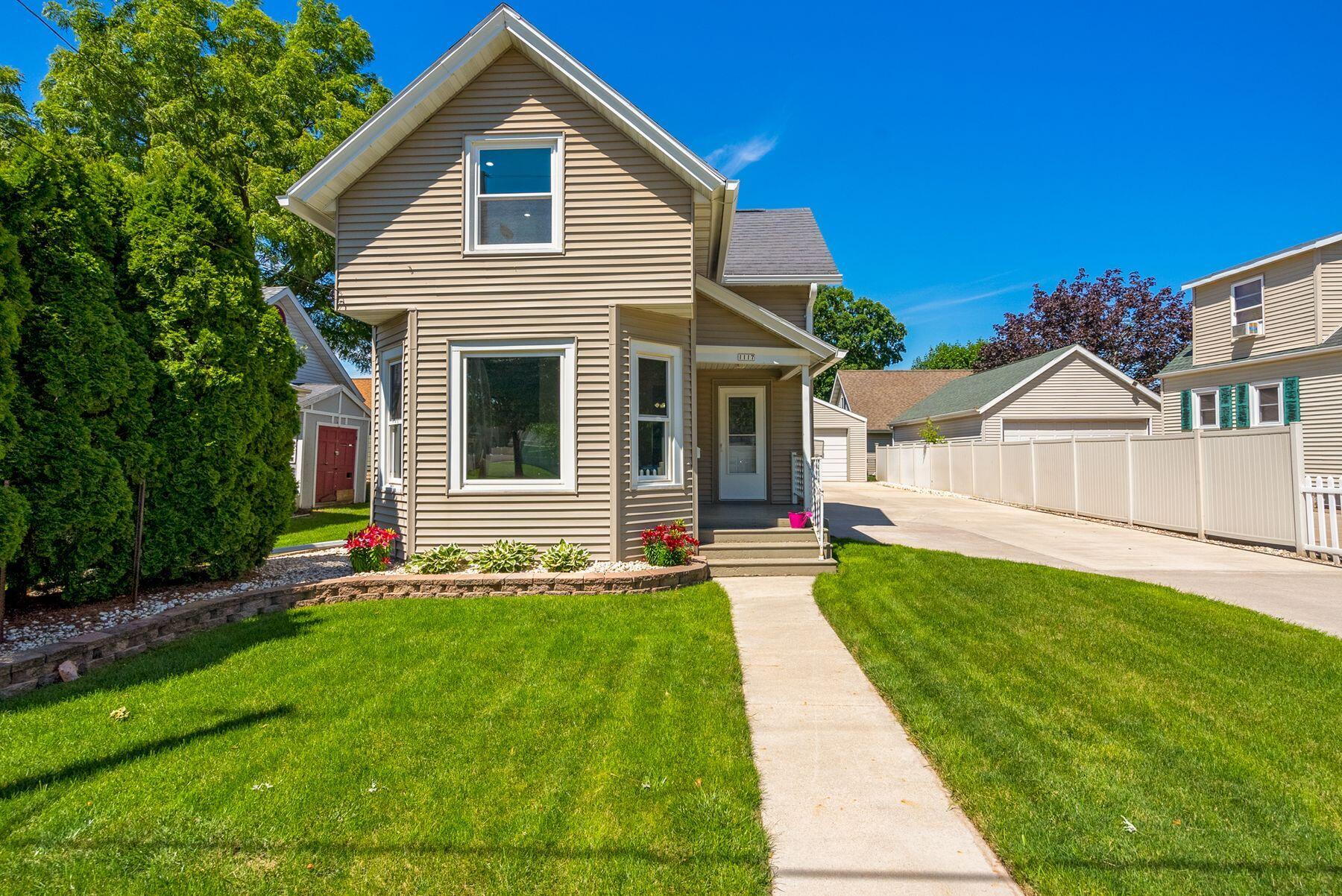 Tan two-story house with a steep front gable, flower beds, and a white-fenced driveway.