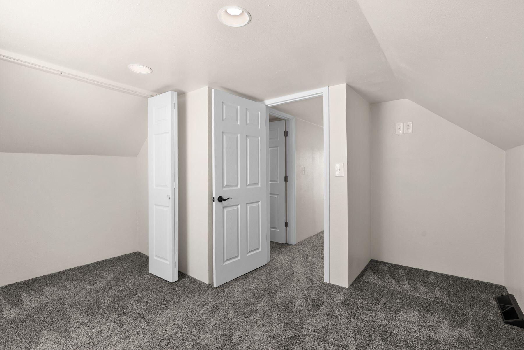 Empty carpeted attic room with a sloped ceiling and open doors to the hallway.