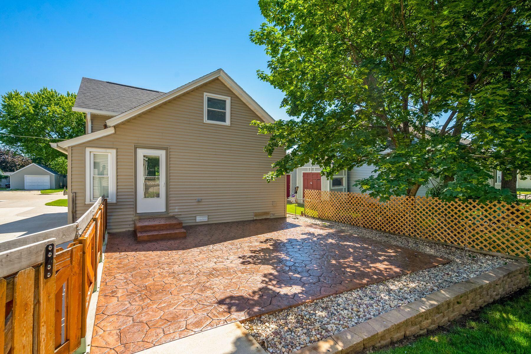 House exterior with a broad stamped-concrete patio, wooden gate, and leafy shade trees.