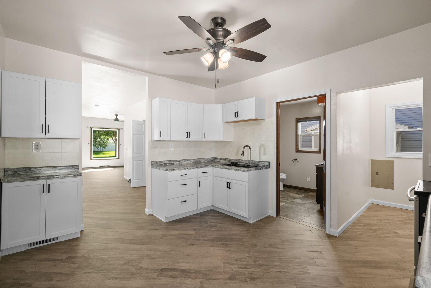 Vacant kitchen with white cabinets, gray countertops, wood-look tile floors, and an adjoining bathroom.