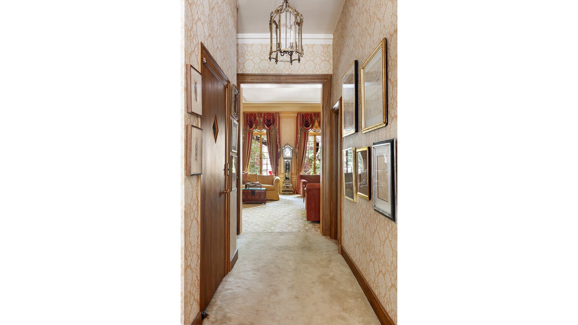 Carpeted hallway with patterned wallpaper opening into a living room with tall windows and red drapes.