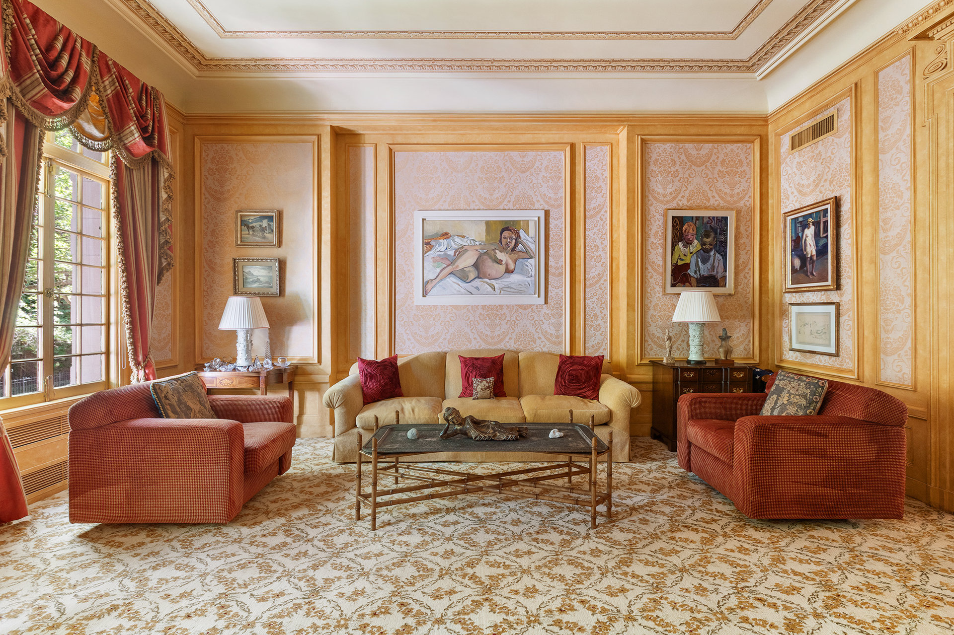 Living room with ornate golden paneling, a cream sofa, red armchairs, and tall draped windows.