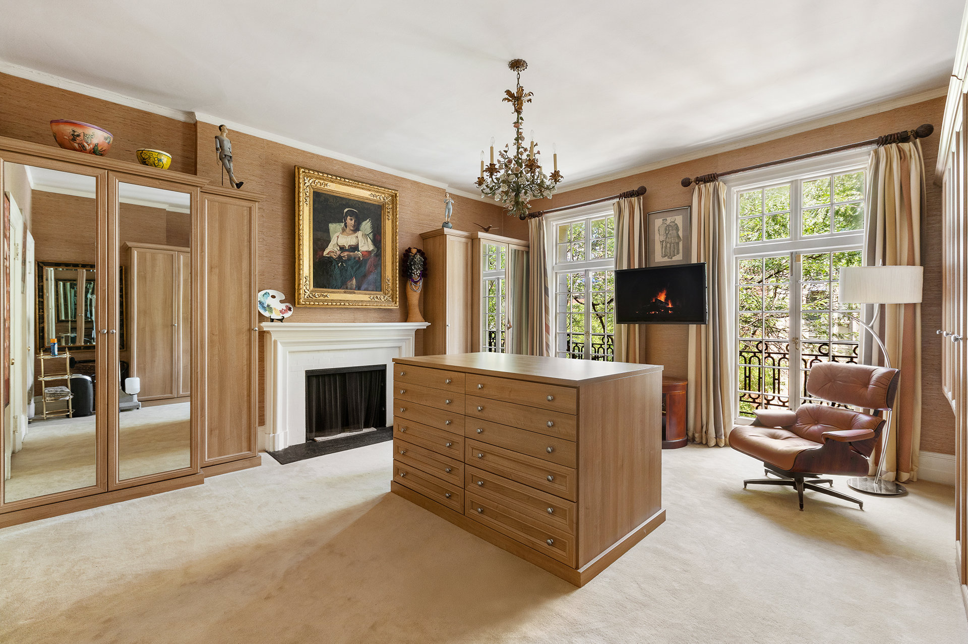 Furnished bedroom with mirrored wardrobes, a fireplace, and tall windows overlooking leafy greenery.