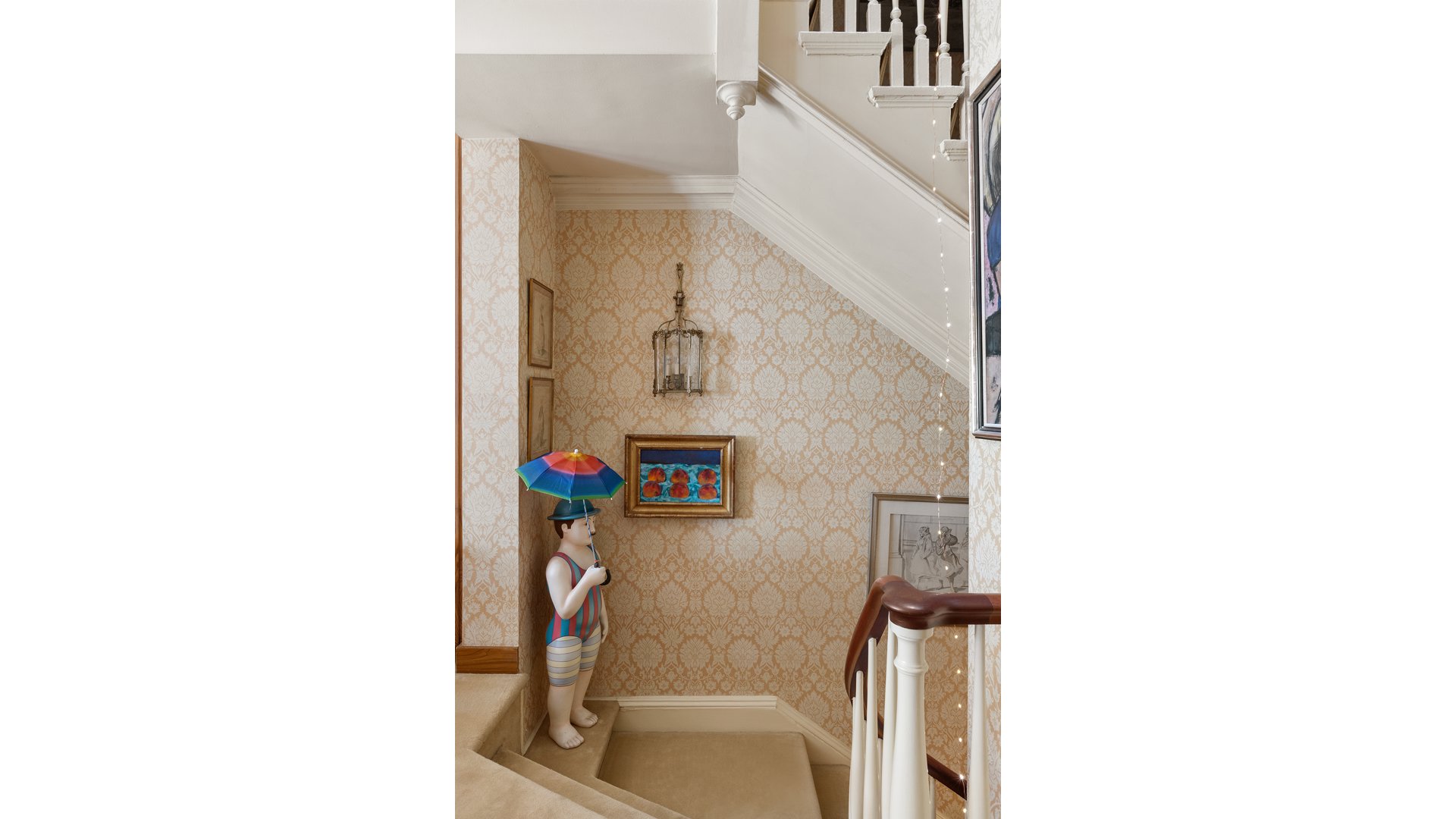 Carpeted staircase with patterned wallpaper, framed art, and a mannequin holding a colorful umbrella.