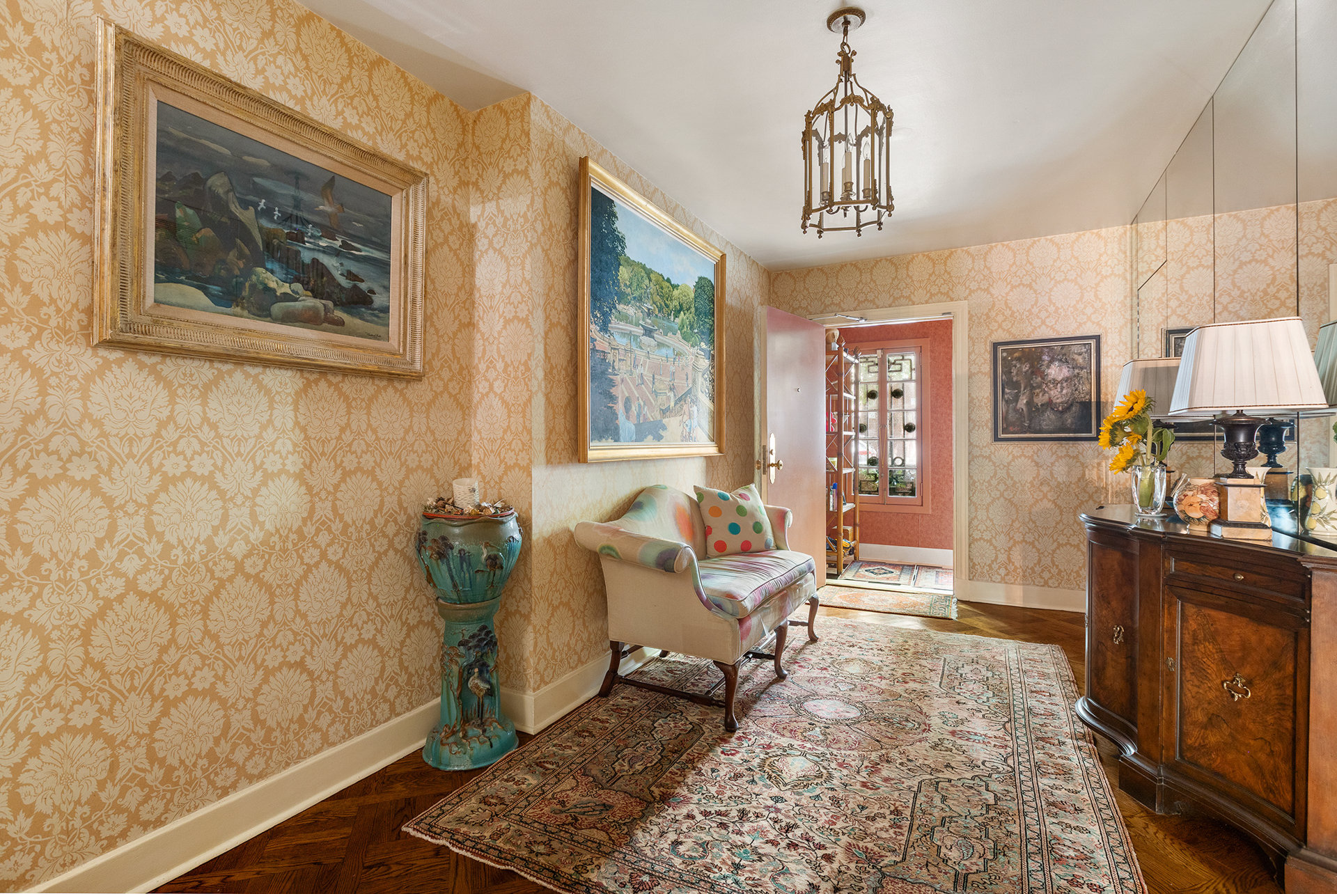 Furnished sitting room with ornate gold wallpaper, framed paintings, and layered patterned rugs.