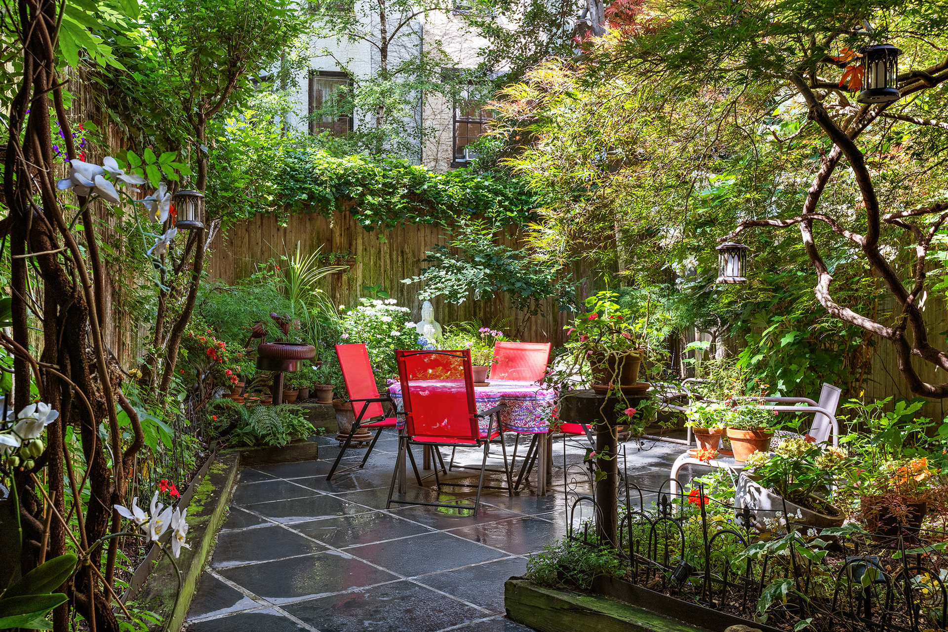 Secluded garden patio with red chairs, a stone terrace, and abundant greenery.