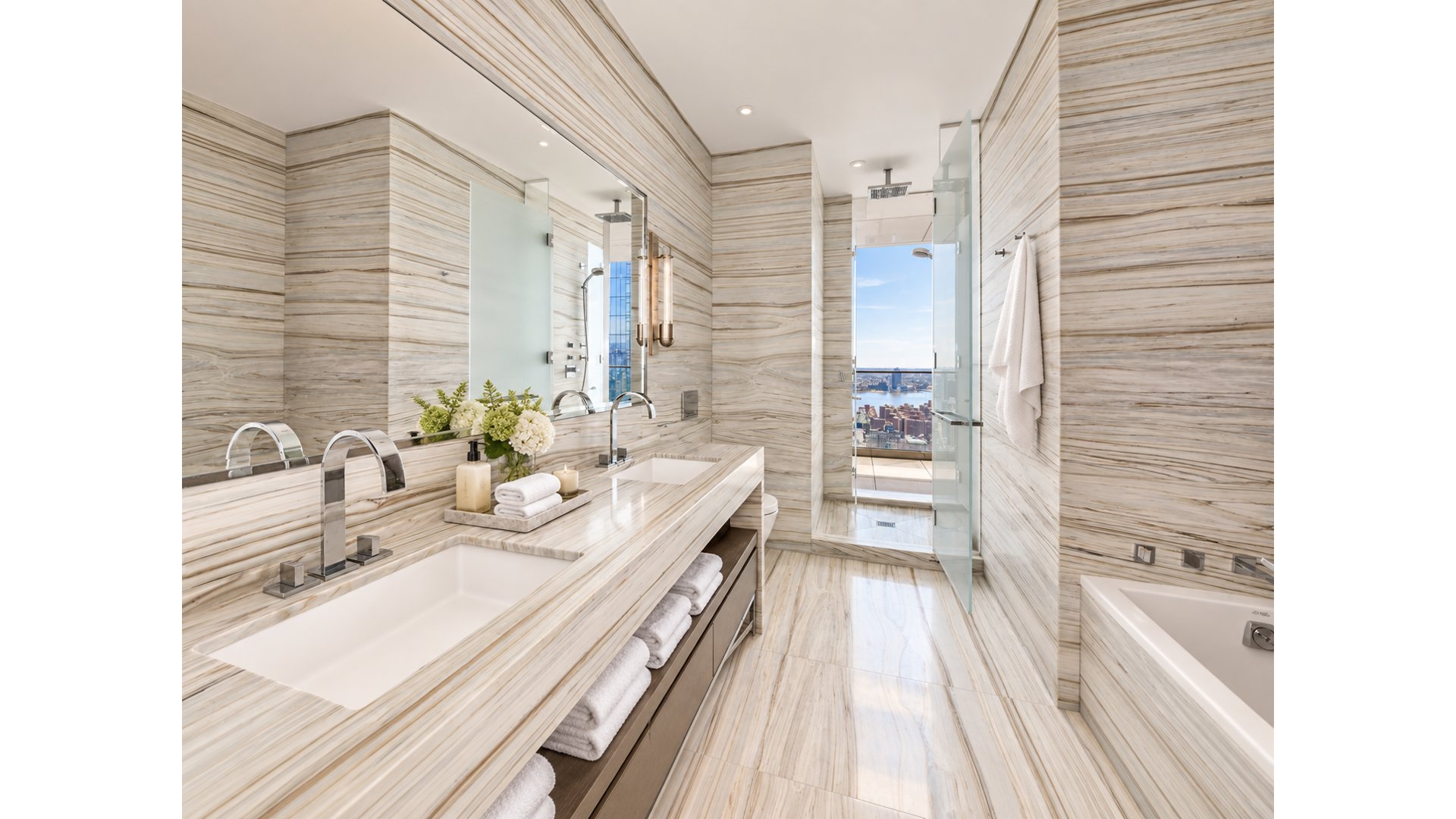 Luxury bathroom with a veined stone double vanity, glass shower, bathtub, and city-and-water view.