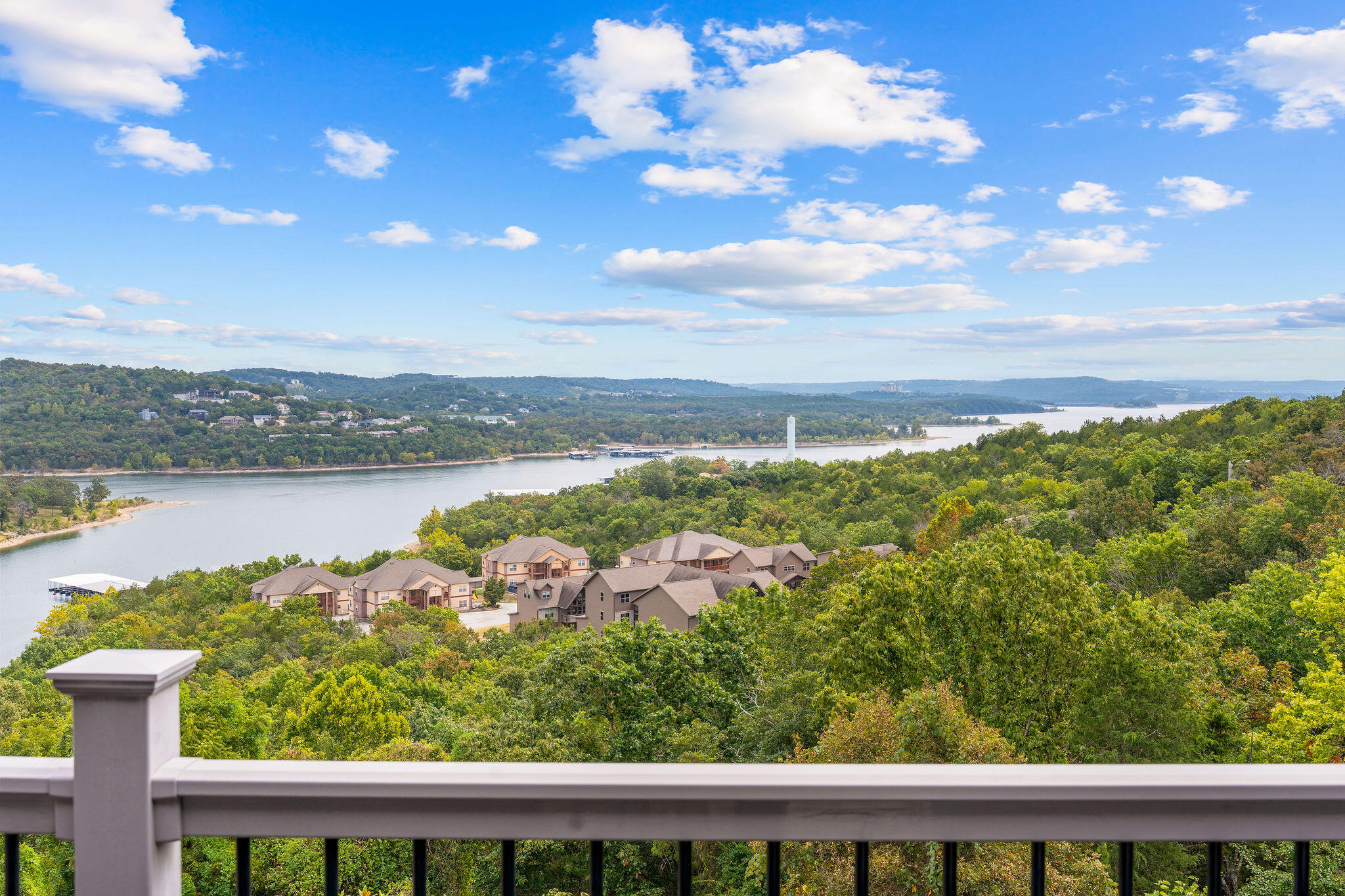 Panoramic lake and wooded hillside view from a balcony, with homes nestled among the trees.