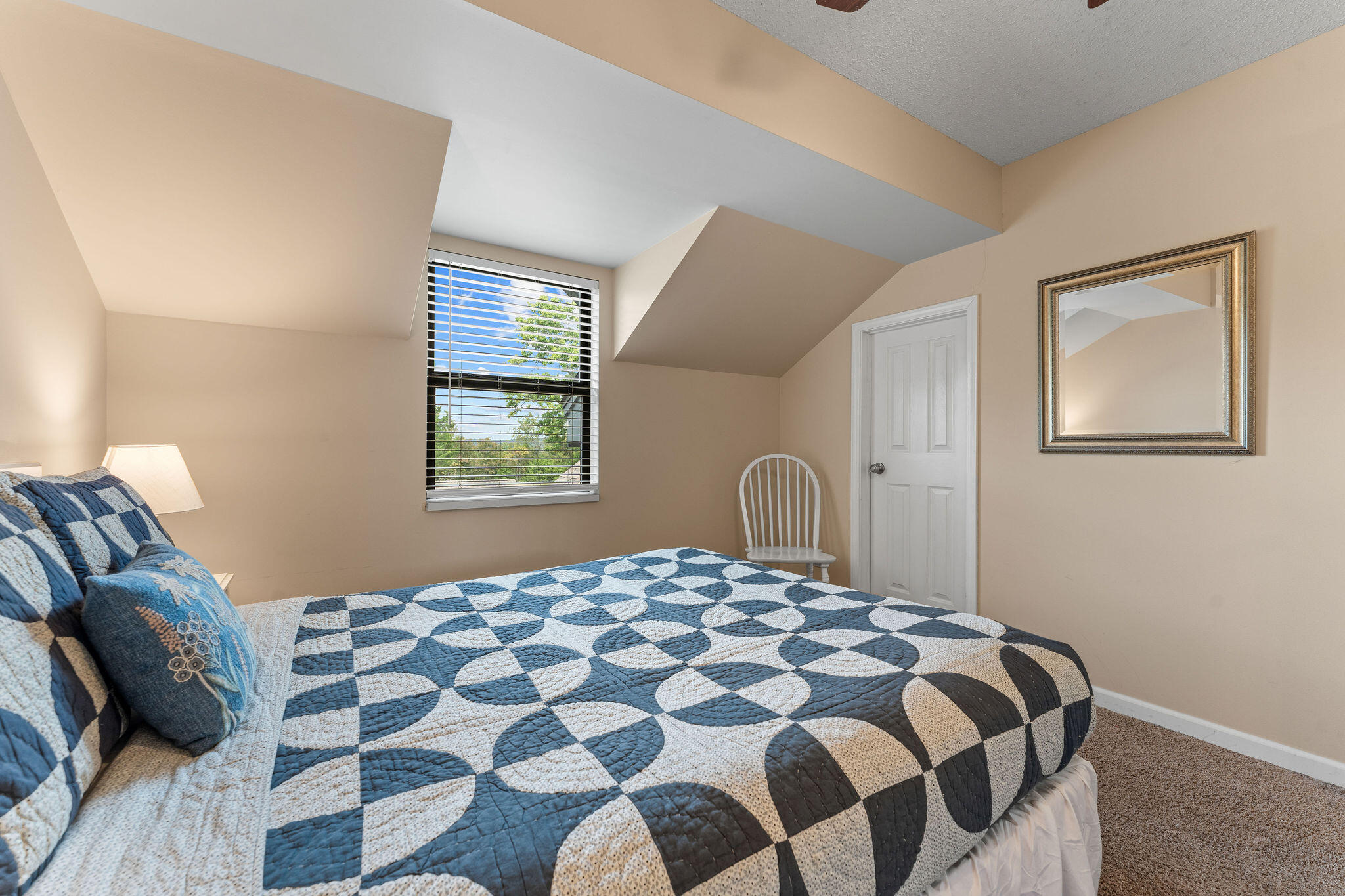 Bedroom with a blue geometric quilt, sloped ceiling, and window overlooking trees.