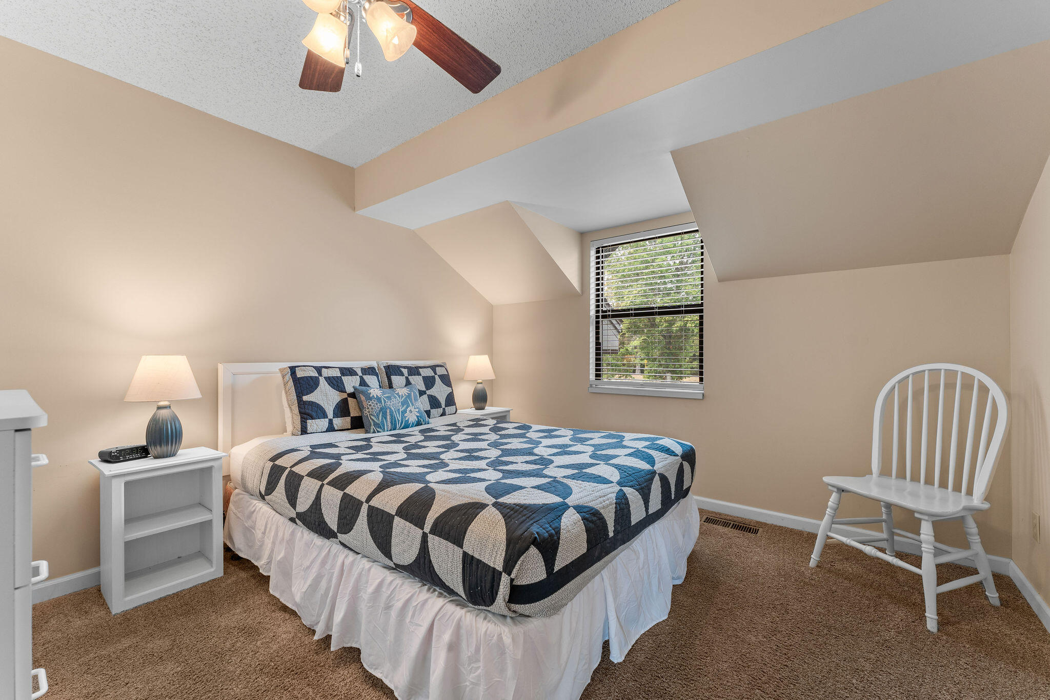 Bedroom with a geometric blue-and-gray quilt, bedside lamps, and a white spindle-back chair.