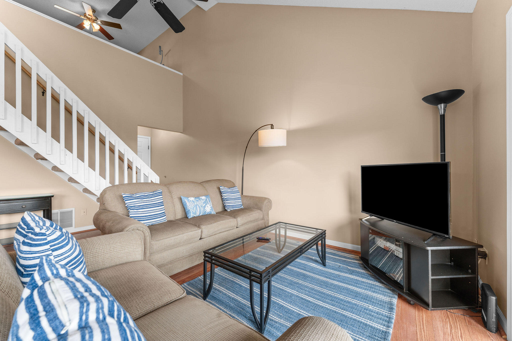 Living room with a beige sectional, blue striped rug, glass coffee table, and television beneath a high ceiling.