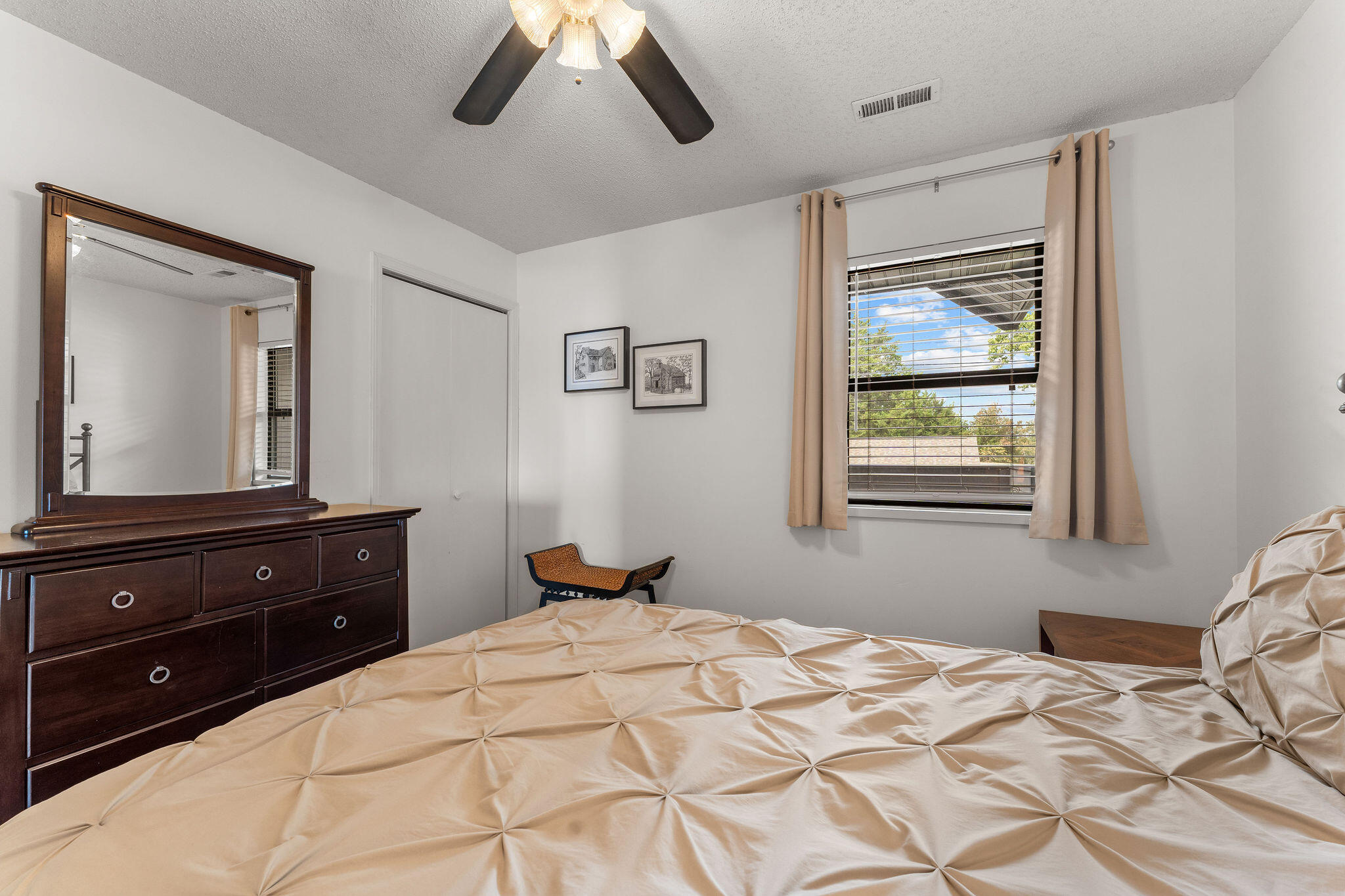 Bedroom with a beige quilted bed, dark wood dresser and mirror, and curtained window.