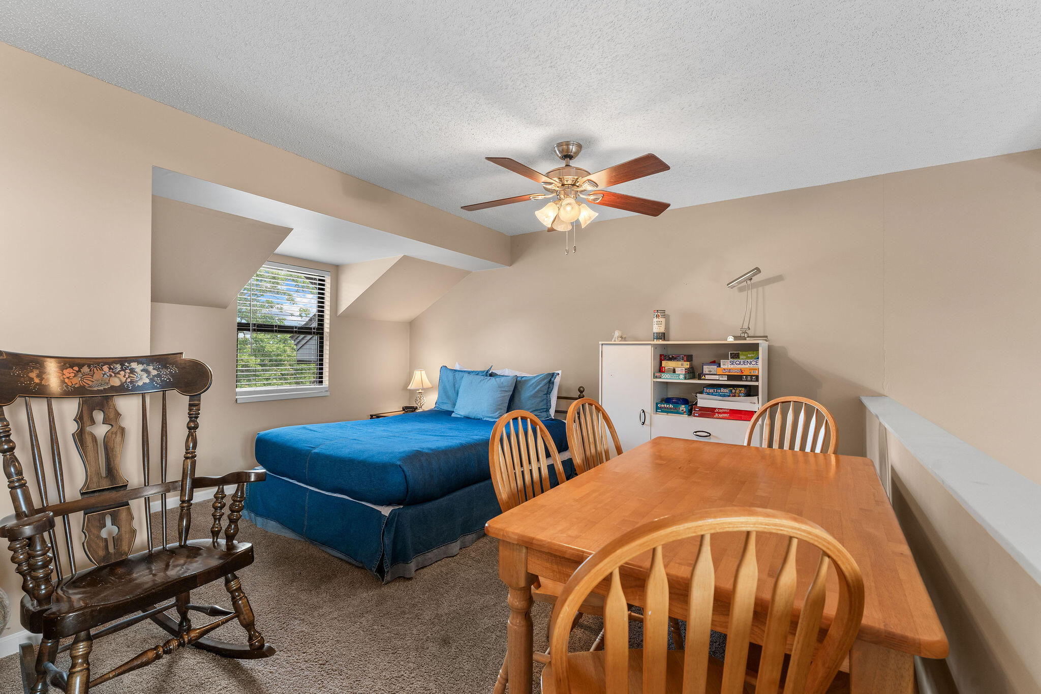 Bedroom with a blue bed, wooden dining table, rocking chair, and board-game shelf.