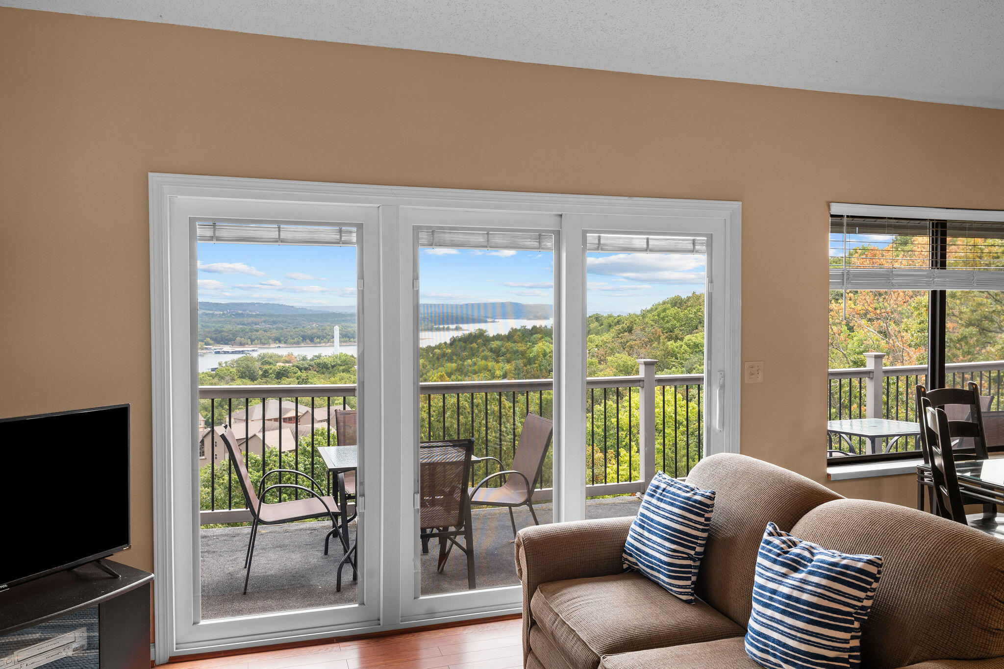 Living room with balcony doors overlooking wooded hills and water, furnished with a sofa and striped pillows.