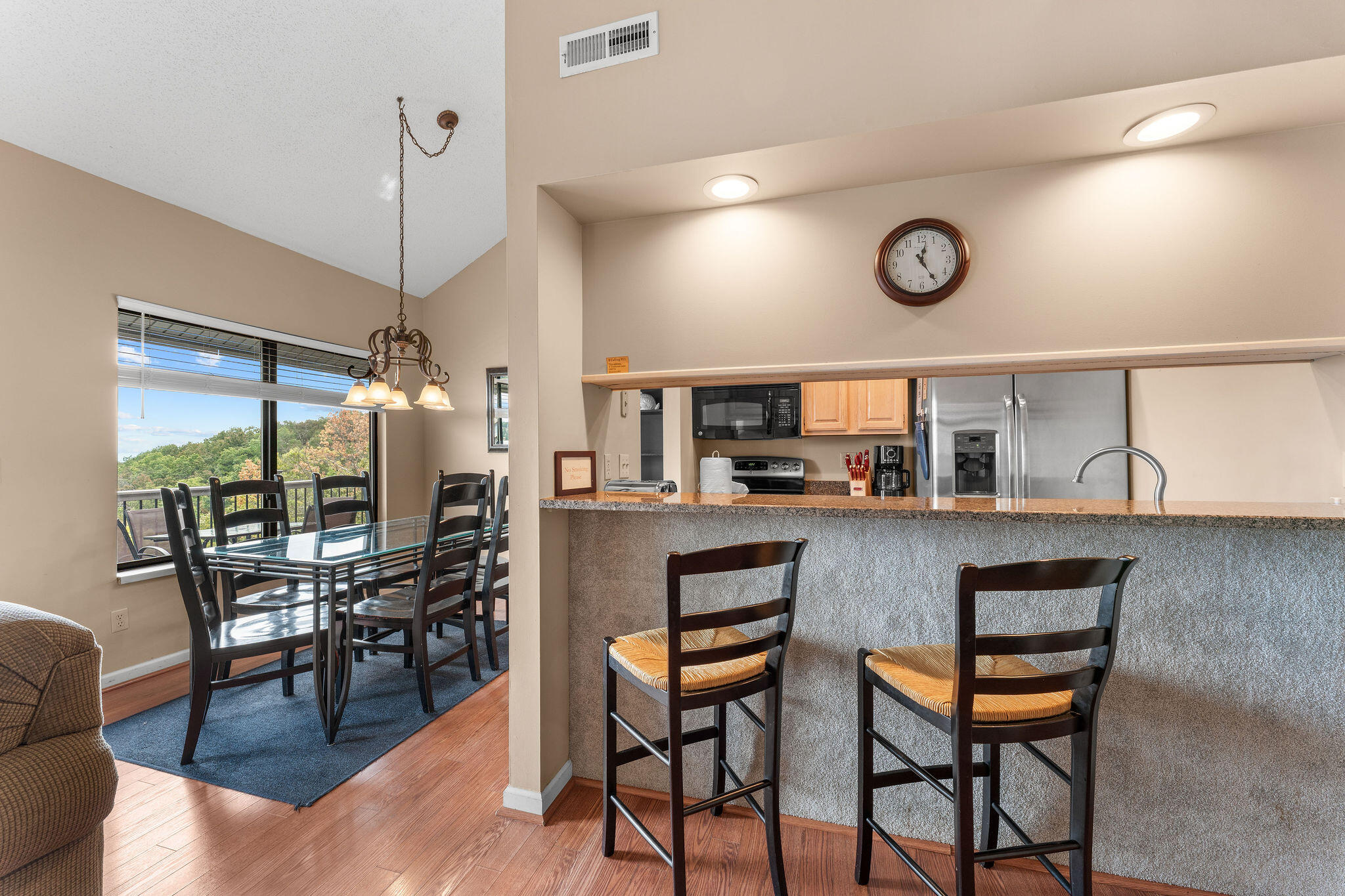 Open kitchen and dining area with a breakfast bar, glass table, and tree views through a large window.