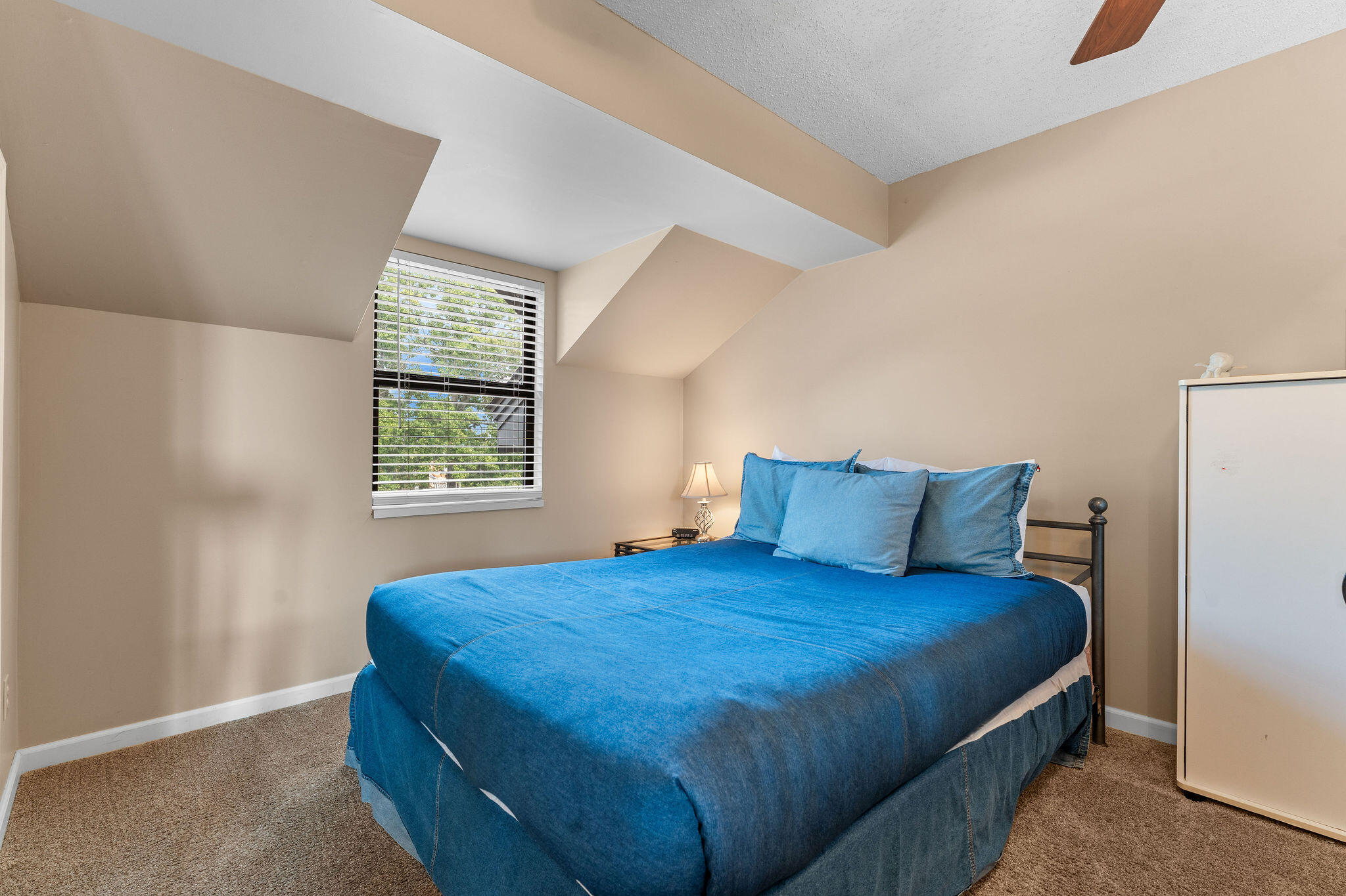 Bedroom with a large blue-covered bed beneath an angled ceiling and a window with white blinds.