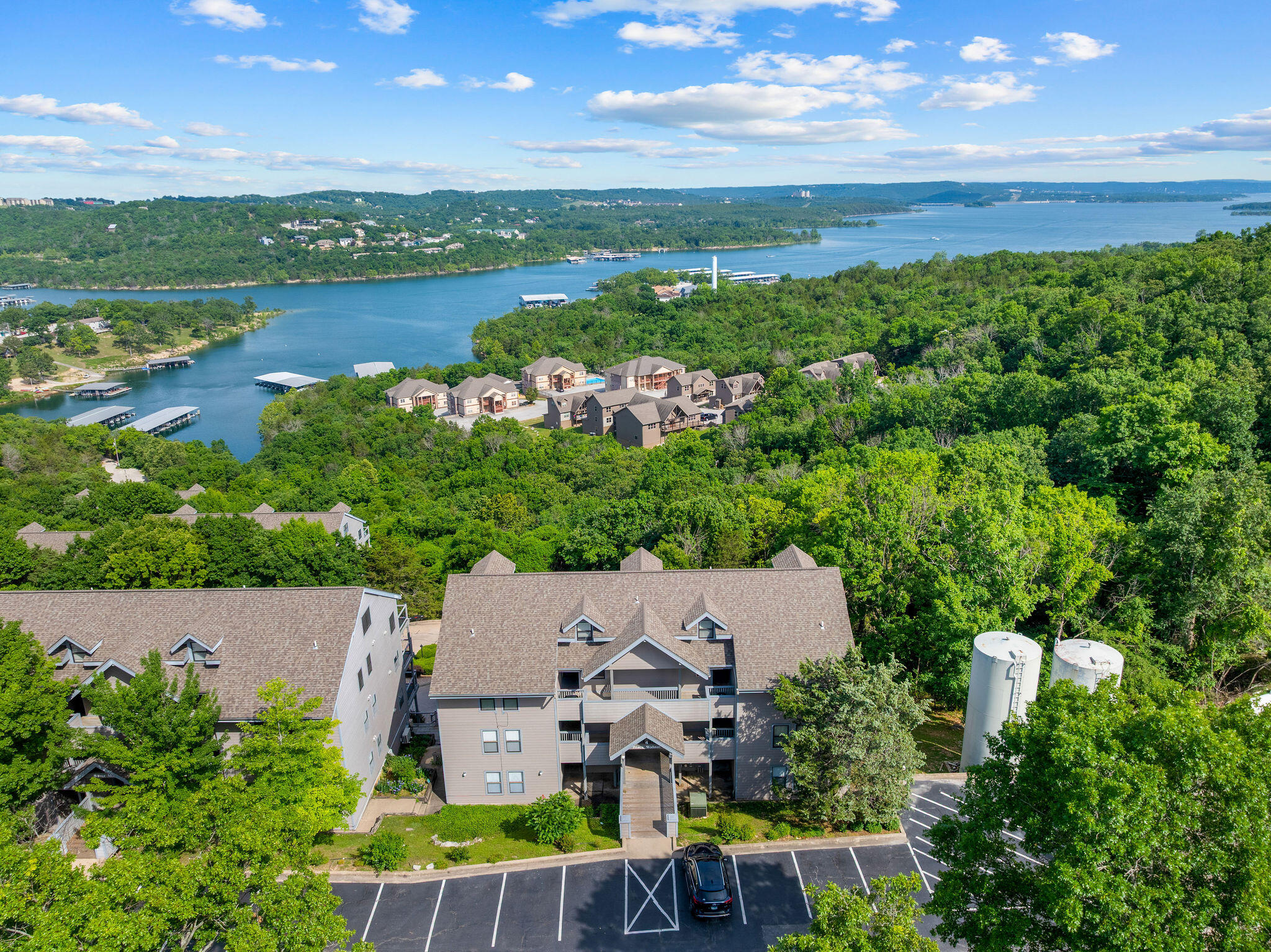 Aerial view of a wooded condominium complex beside a lake, with forested shorelines in the distance.