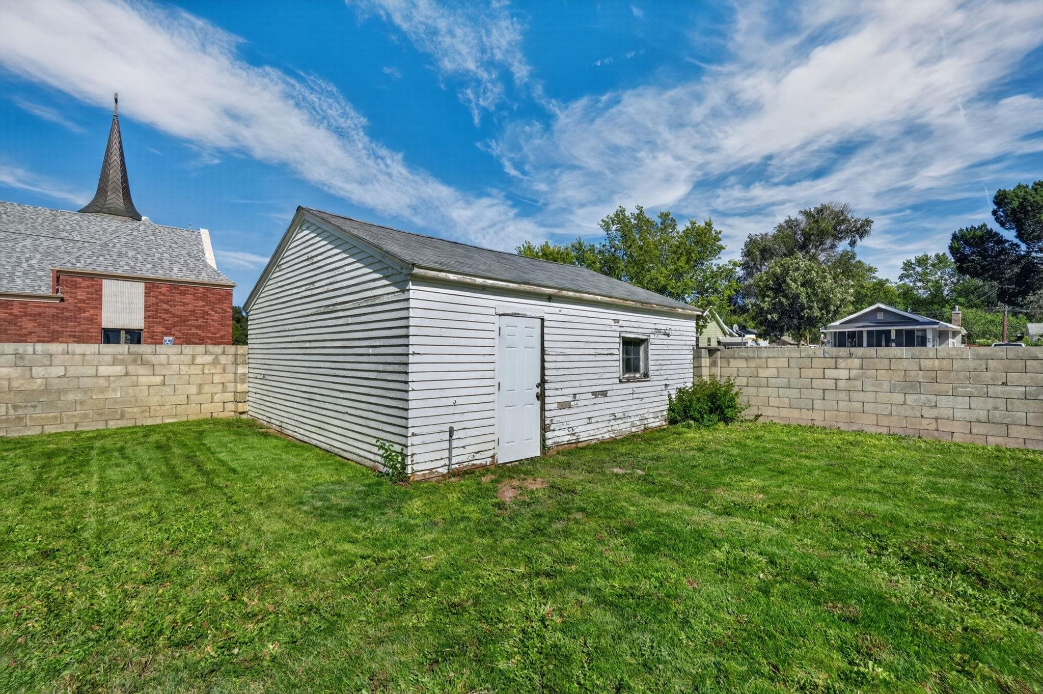 Weathered white outbuilding with a gabled roof and white door in a block-walled backyard.