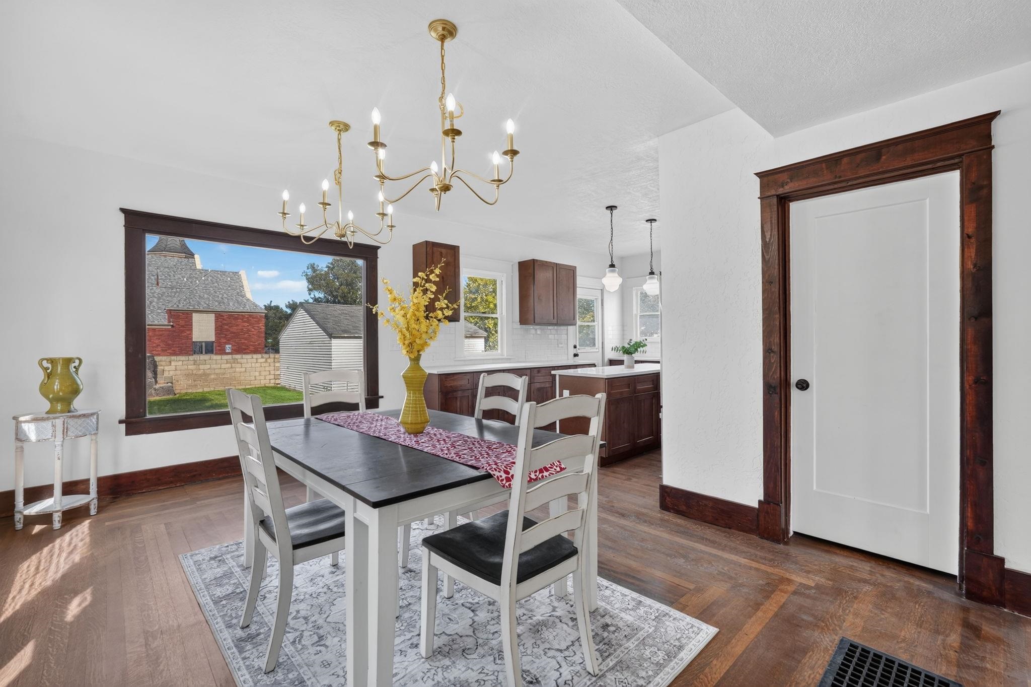 Dining room with a dark table, white chairs, brass chandeliers, and an adjoining kitchen.