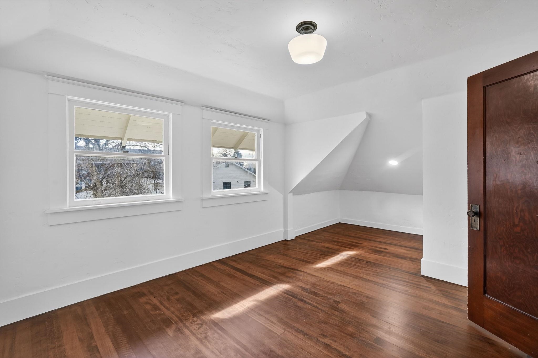 Empty room with polished wood floors, two windows, and a sloped ceiling alcove.