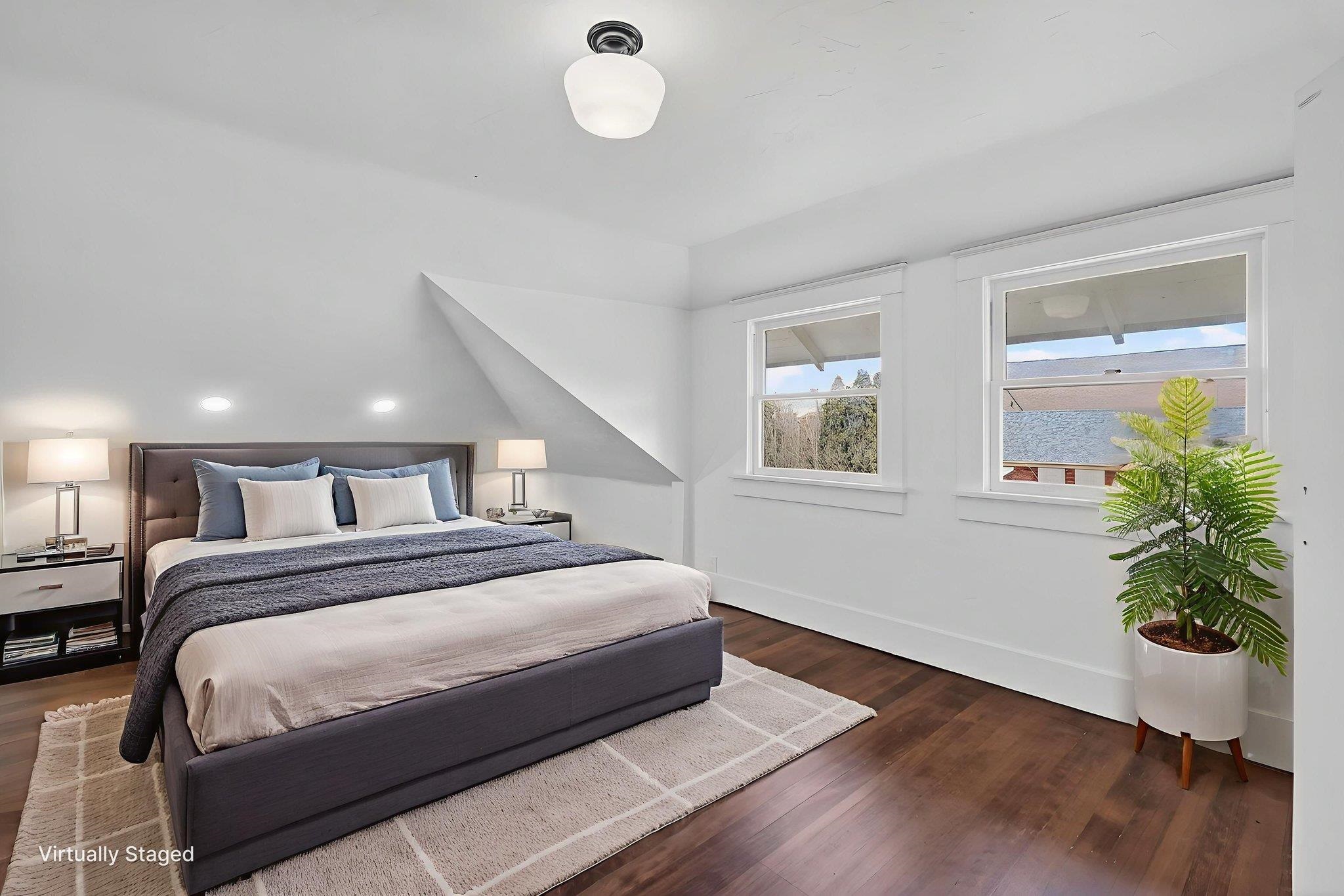 Virtually staged bedroom with a large bed beneath a sloped ceiling and two windows.